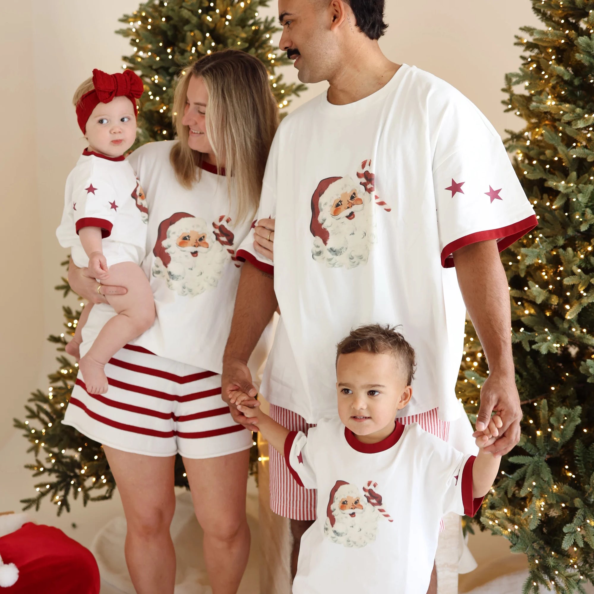 Family of four in matching vintage Santa print tees and red stripe shorts between lit Christmas trees