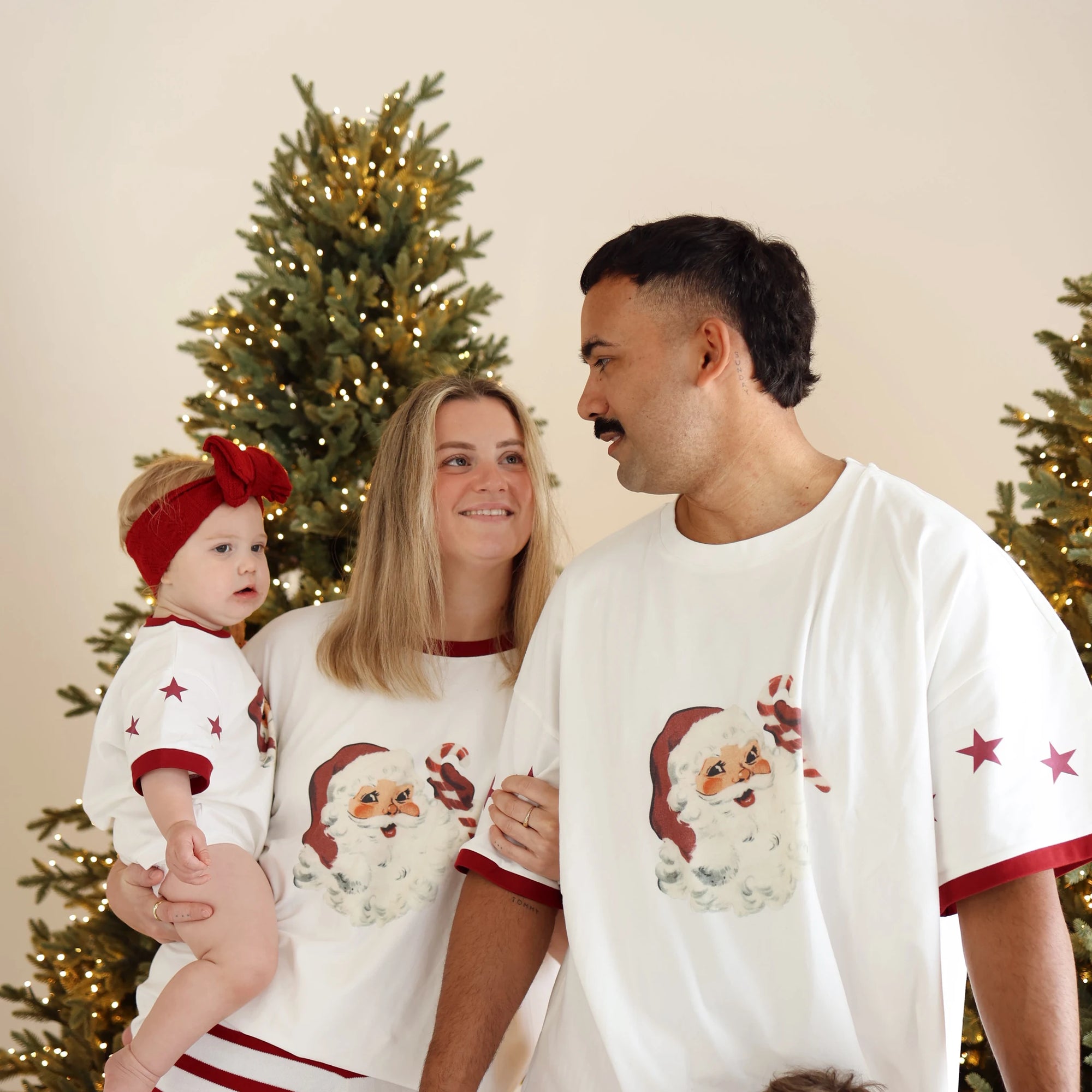 Family in matching vintage Santa print tee pyjamas, mum holding a baby in a red bow headband, between lit Christmas trees