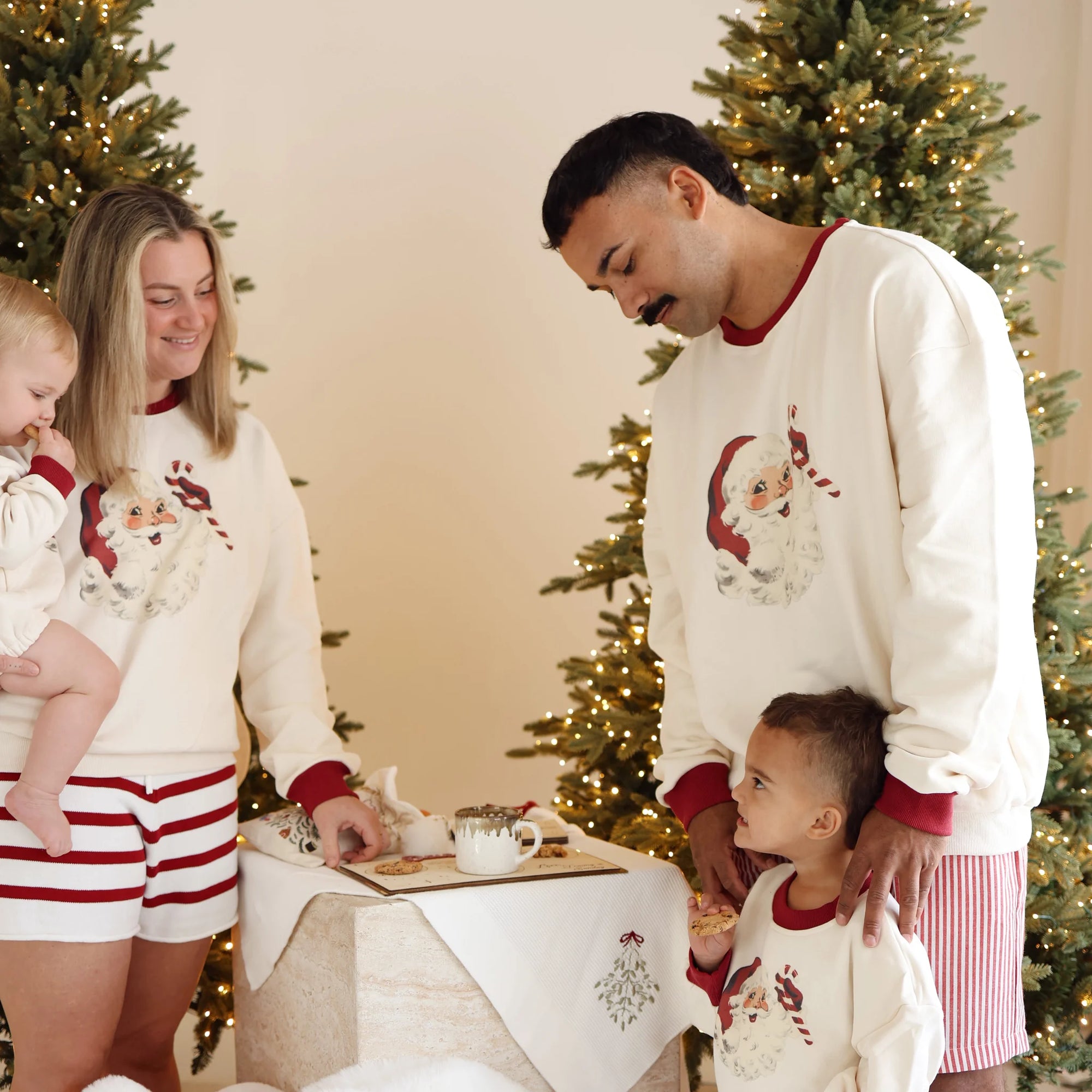 Family in matching vintage Santa print jumpers setting out milk and cookies between lit Christmas trees