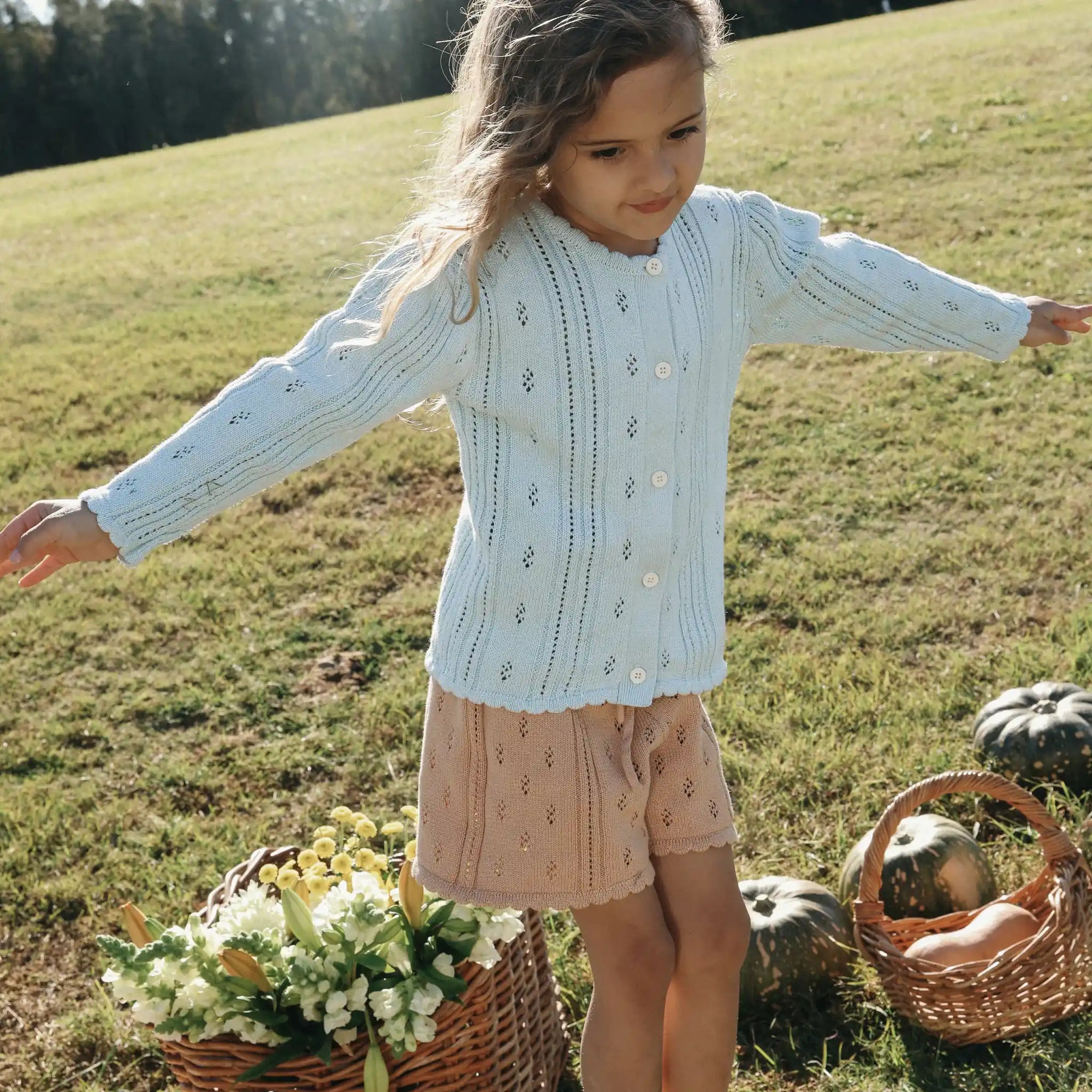 Girl in a pale blue pointelle knit cardigan and apricot shorts with arms outstretched beside flower baskets