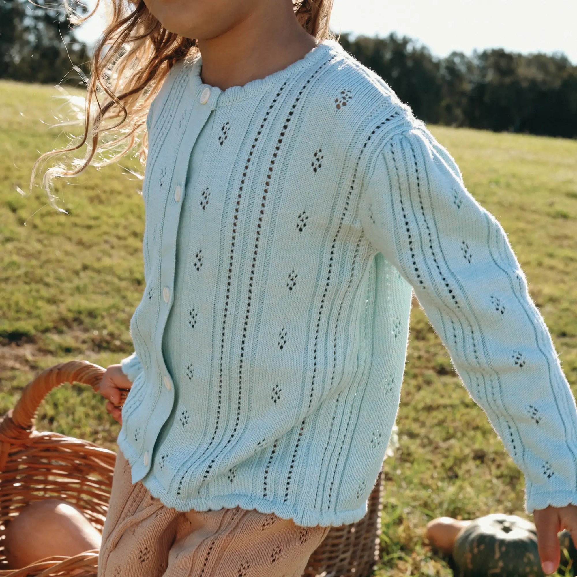 Girl in a pale blue pointelle knit cardigan and cream pointelle shorts beside a wicker basket