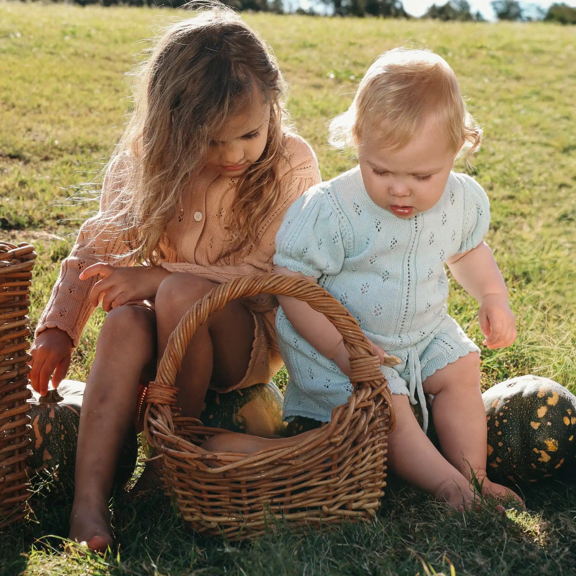 Girl and baby in apricot and pale blue pointelle knit sets sitting beside a basket and pumpkins