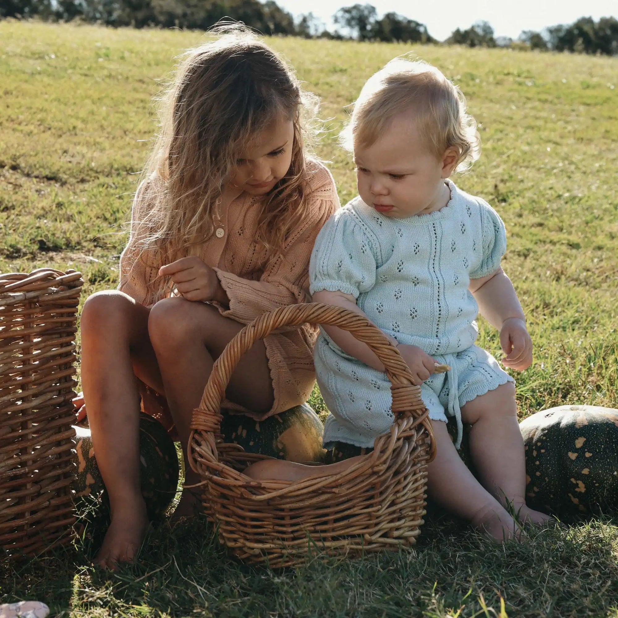 Girl in an apricot pointelle set and baby in pale blue pointelle looking into a wicker basket together
