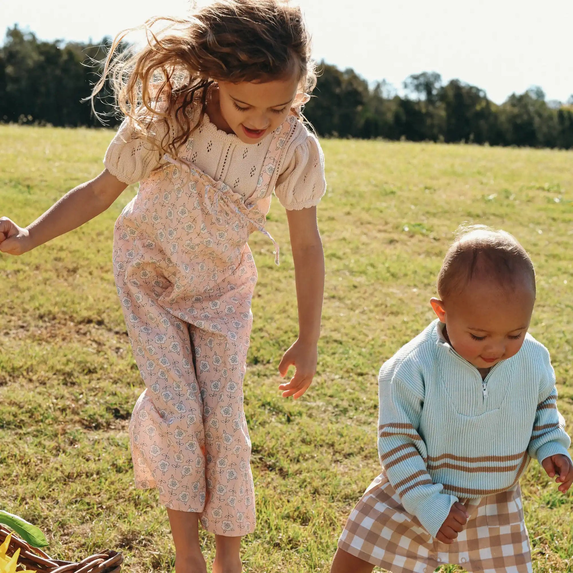Girl in a floral print overall set beside a baby in a pale blue stripe knit jumper and gingham shorts