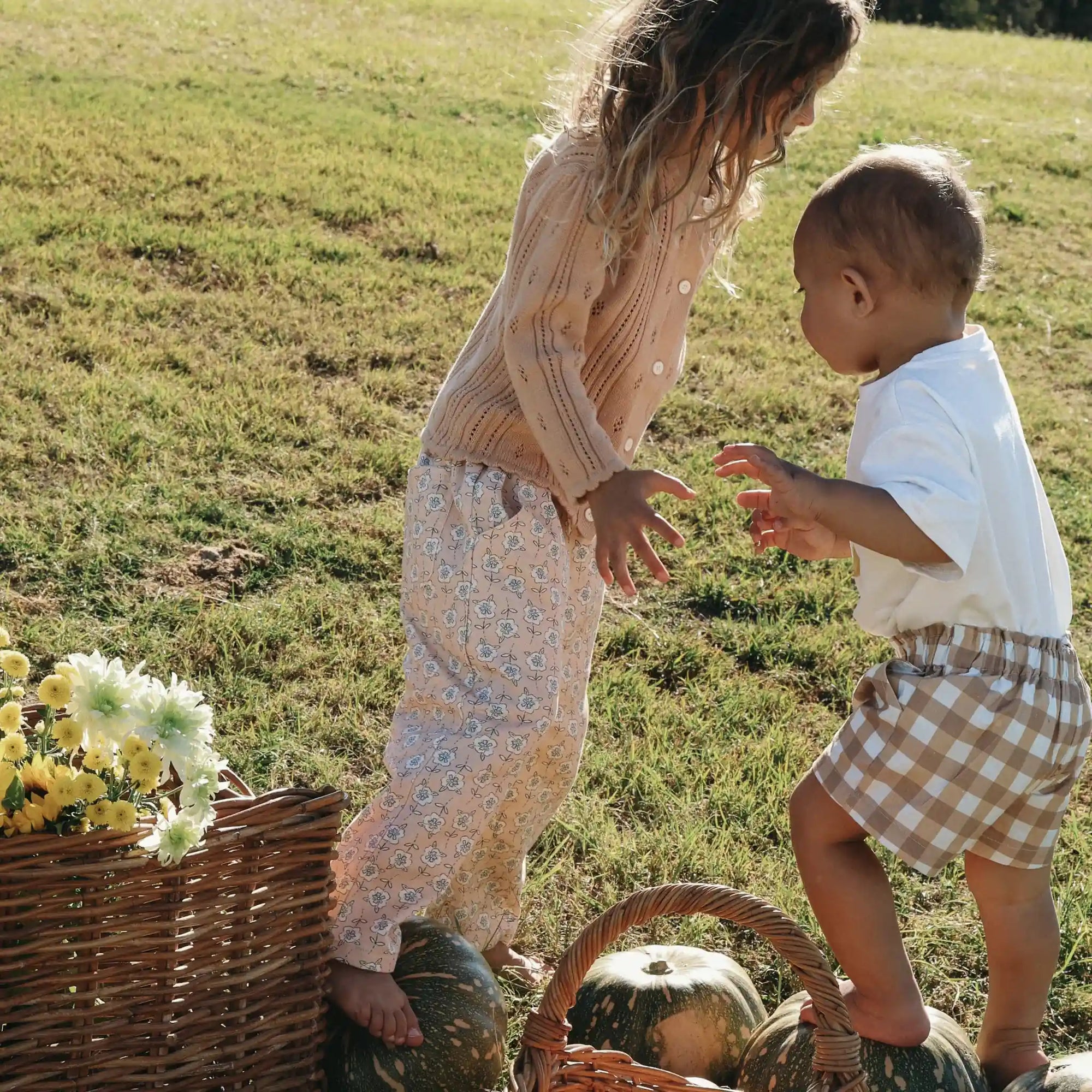Girl in a pointelle cardigan and floral pants reaching for a toddler in a cream tee and gingham shorts