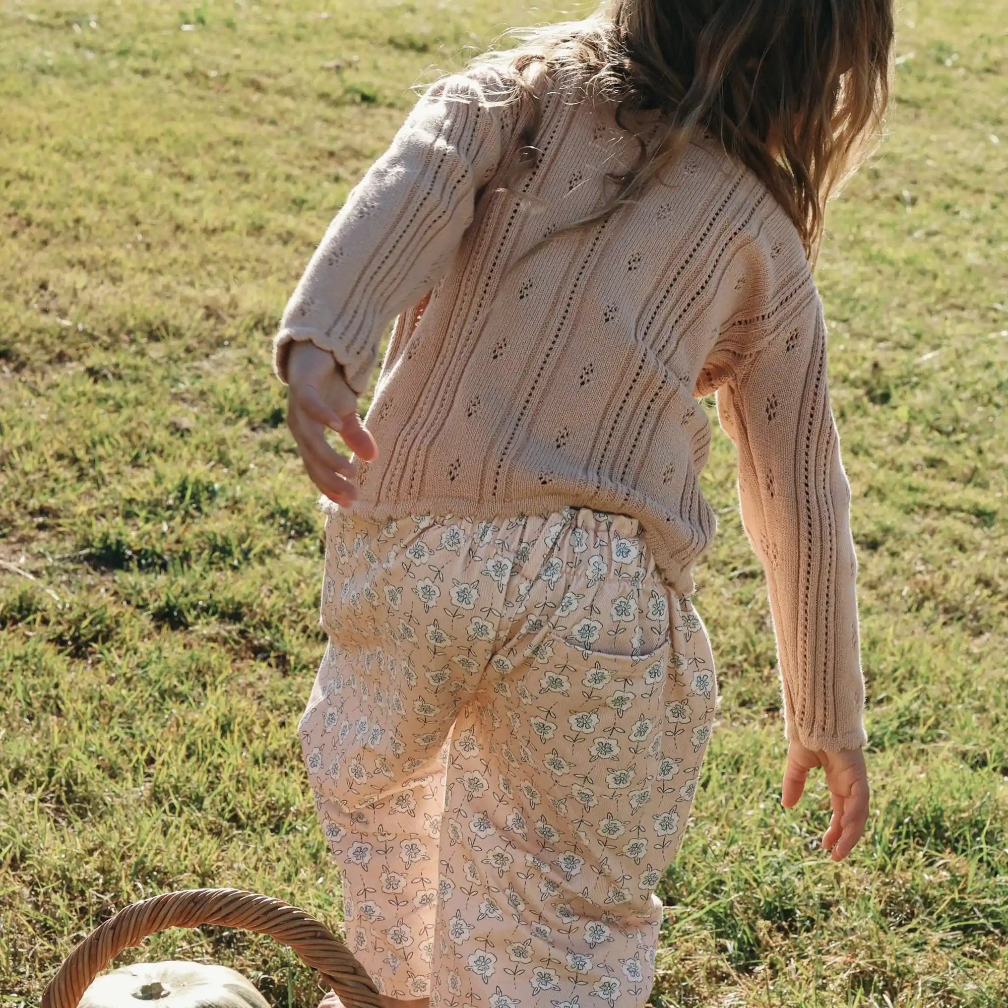 Back view of a girl in a cream pointelle knit cardigan and floral print pants in a sunlit field