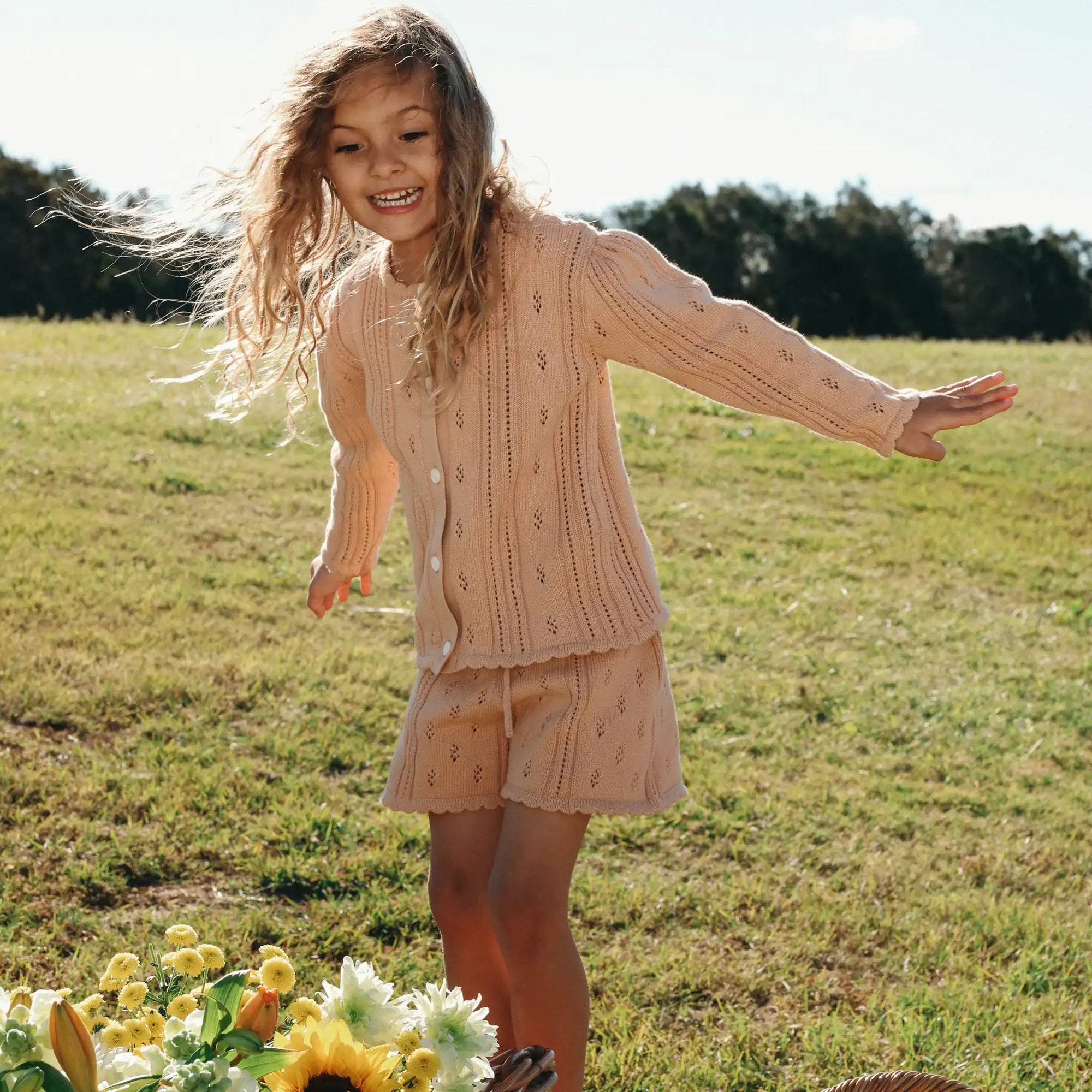 Girl laughing in an apricot pointelle knit cardigan and shorts set beside a basket of flowers