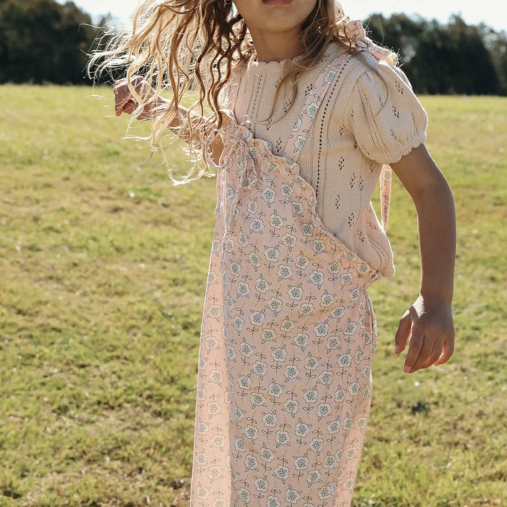 Girl in a cream pointelle knit top and apricot floral print overalls running in a sunlit field