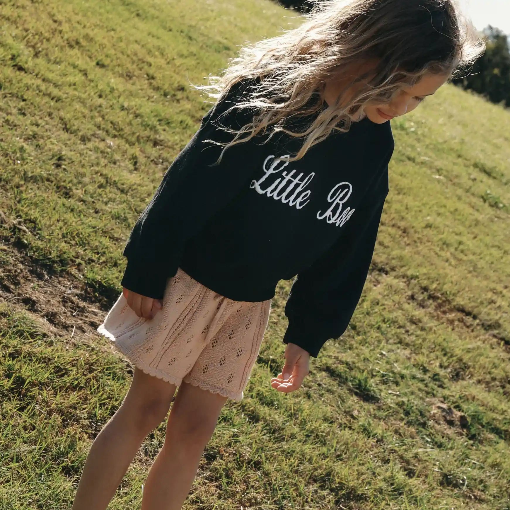 Girl in a black Little Boo embroidered sweatshirt and cream pointelle shorts standing on sunlit grass