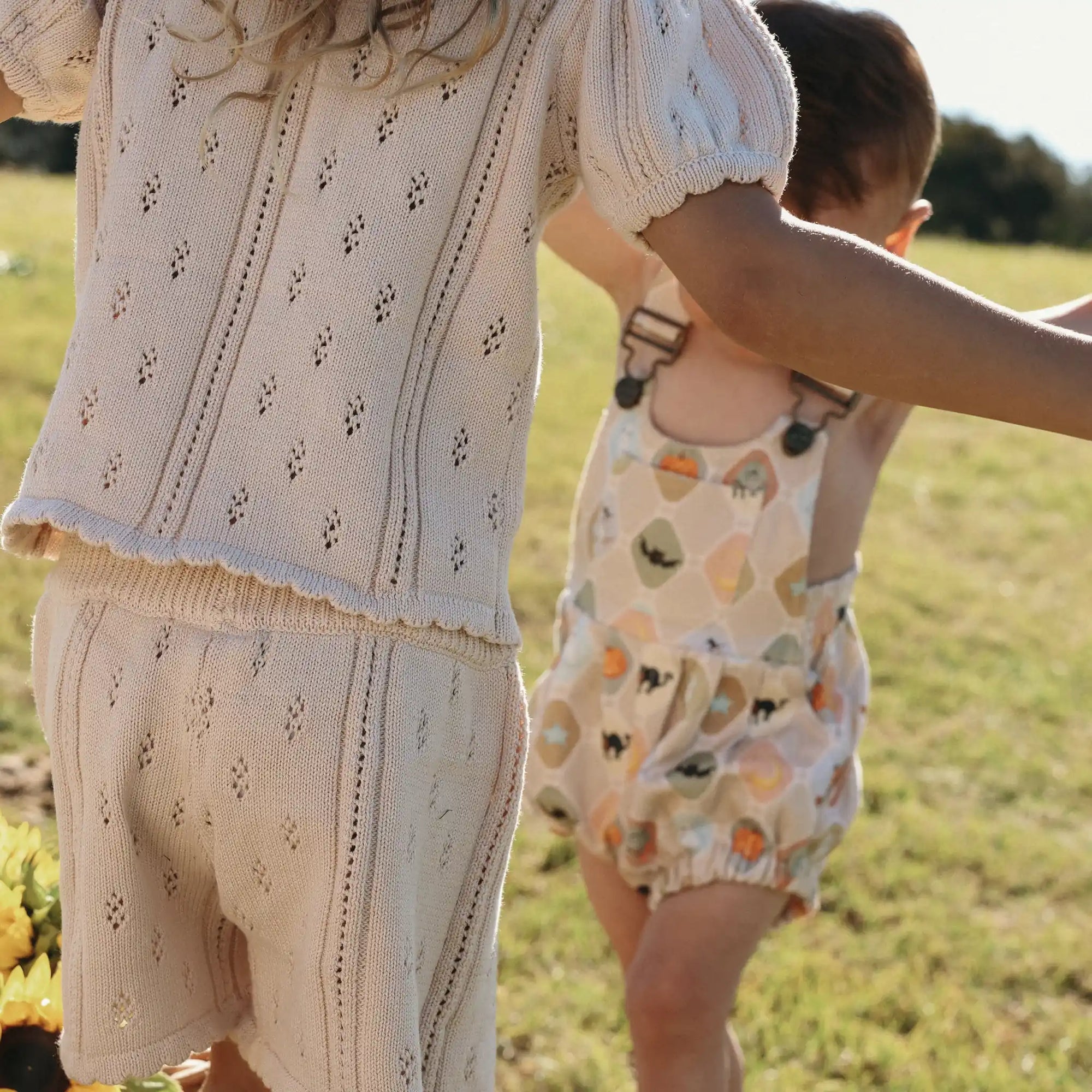 Girl in a cream pointelle knit set holding hands with a baby in a Halloween print overall romper
