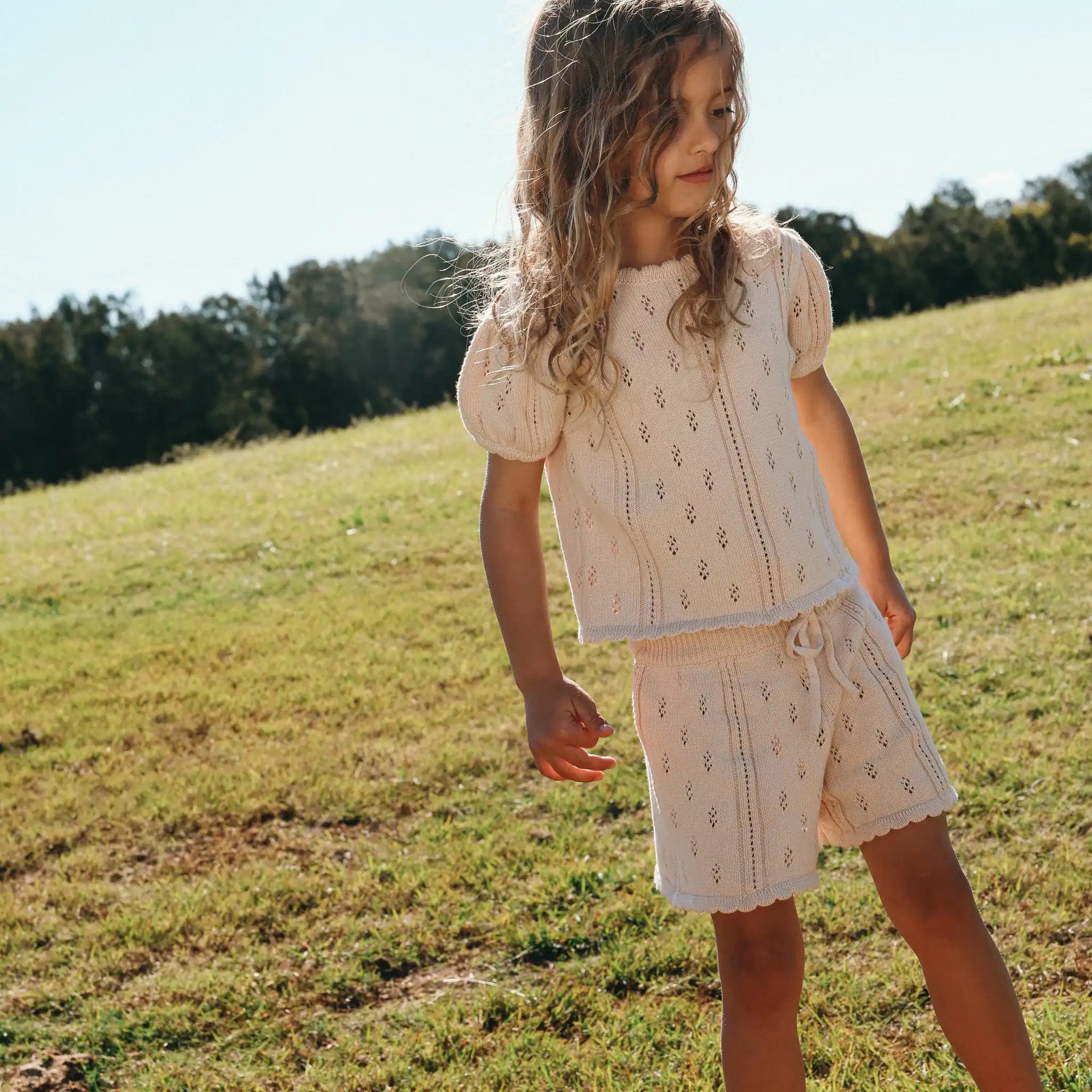 Girl standing in a cream pointelle knit puff sleeve top and drawstring shorts set on a grassy hill