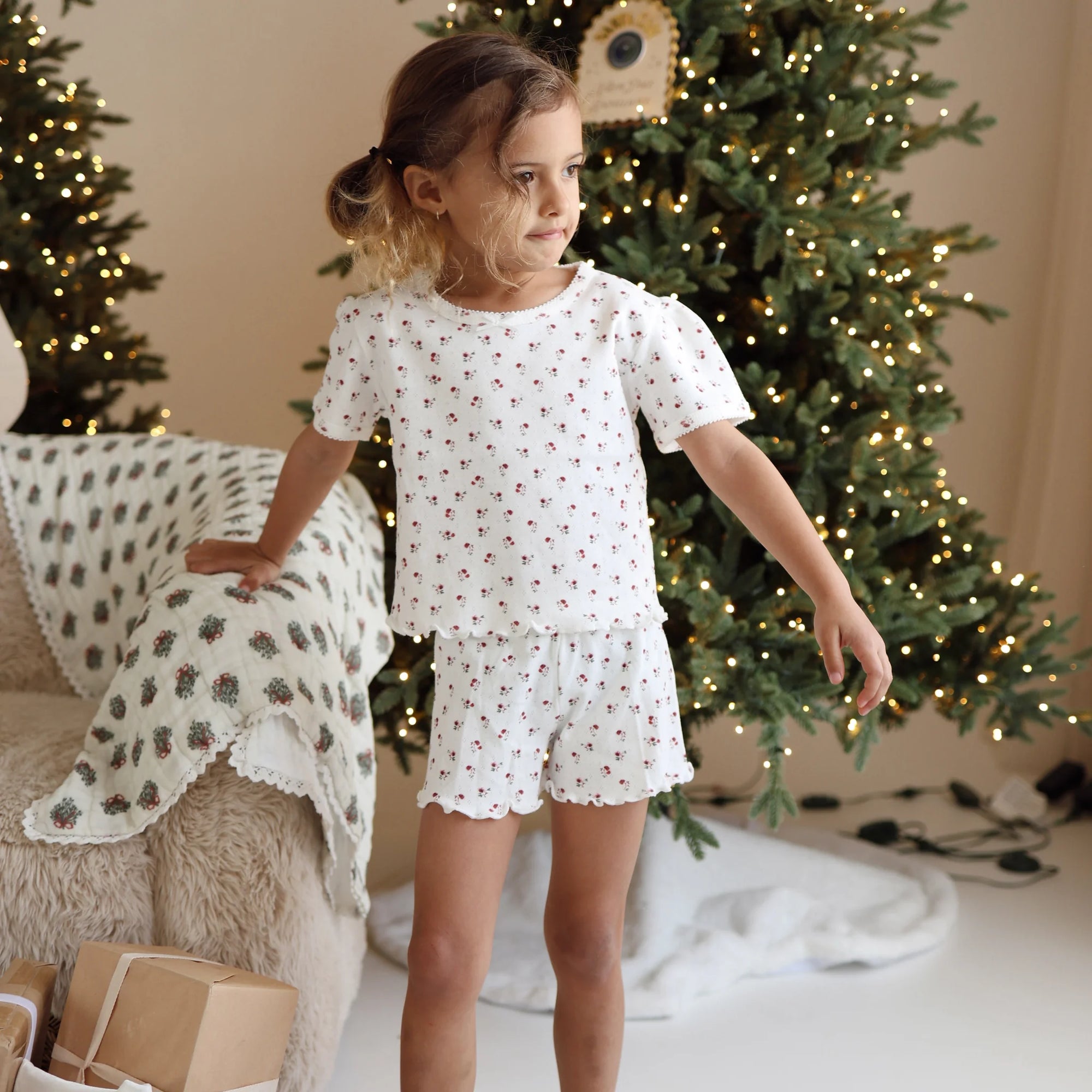 Girl in a cherry print tee and shorts pyjama set standing beside a sheepskin chair and lit Christmas tree