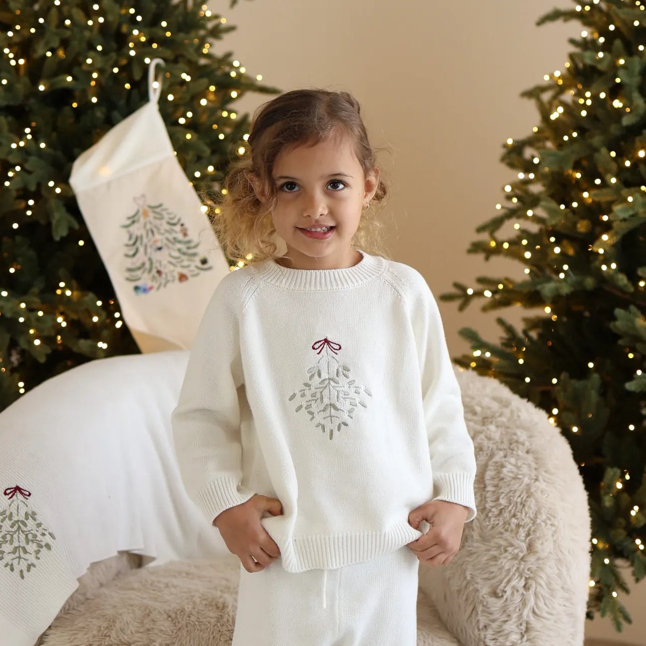 Girl in a cream knit jumper with embroidered mistletoe and matching pants, beside an embroidered Christmas stocking