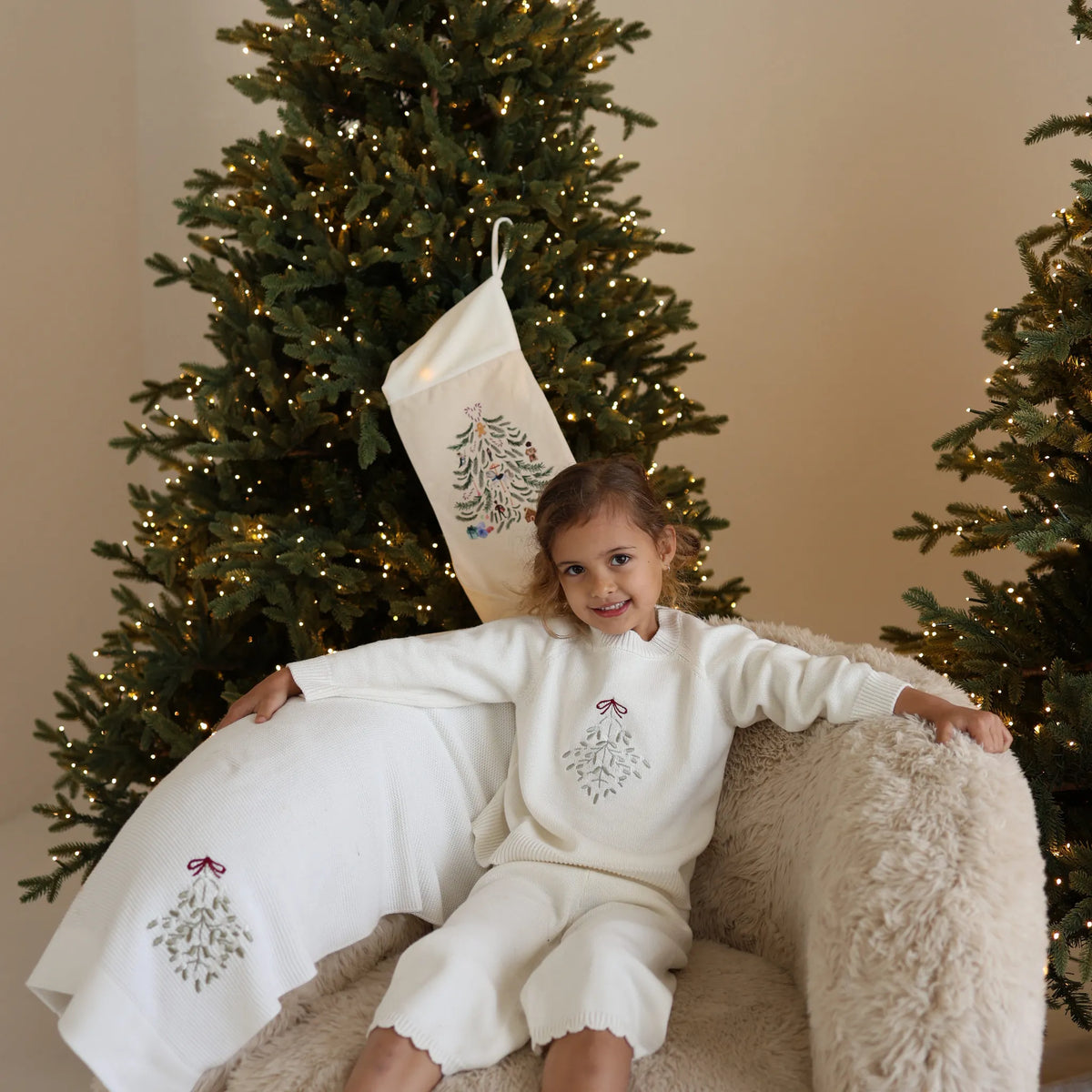 Girl in a cream knit Christmas set with embroidered mistletoe, sitting on a sheepskin chair with a matching blanket and stocking