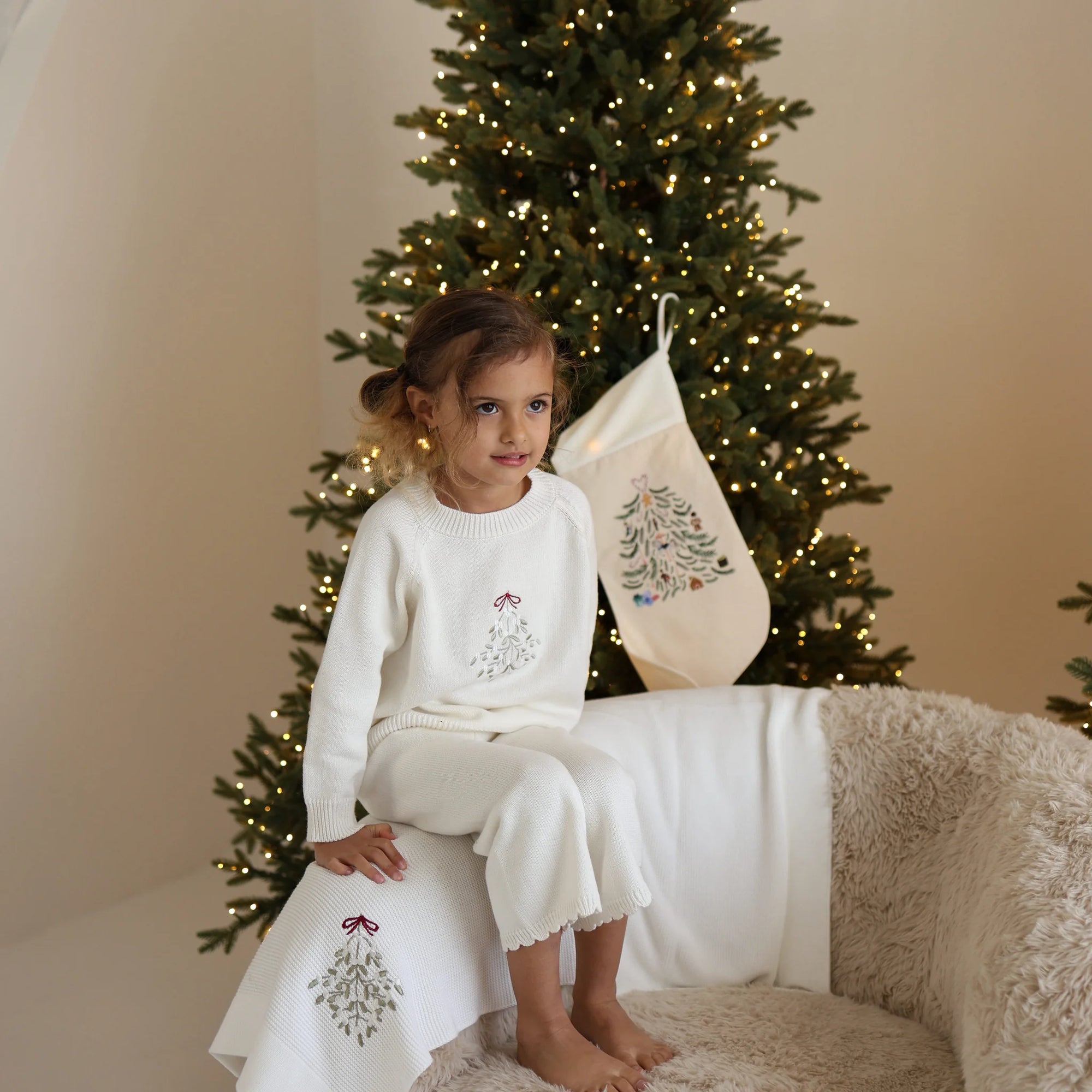 Girl in a cream knit Christmas set with embroidered mistletoe sitting on a sheepskin chair with a matching blanket