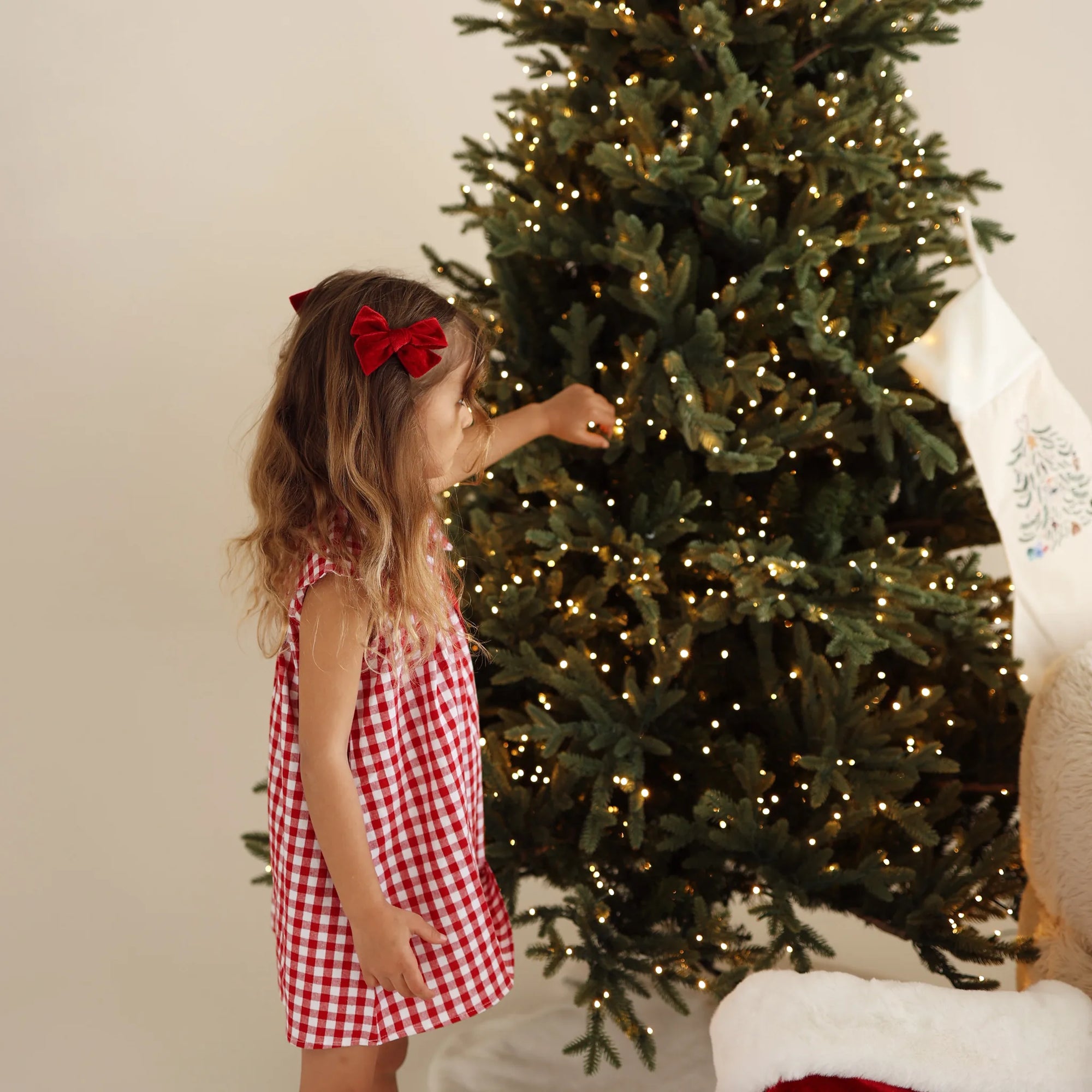 Girl in a red gingham Christmas dress and red velvet hair bow reaching up to decorate a lit Christmas tree