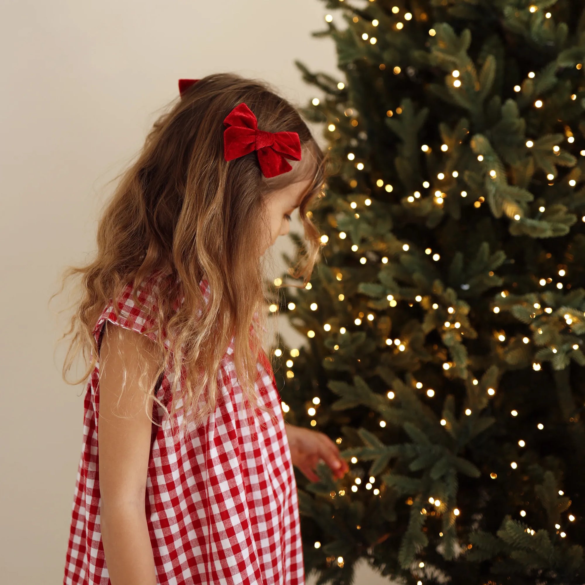 Girl in a red gingham Christmas dress with a red velvet hair bow, looking down beside a lit Christmas tree