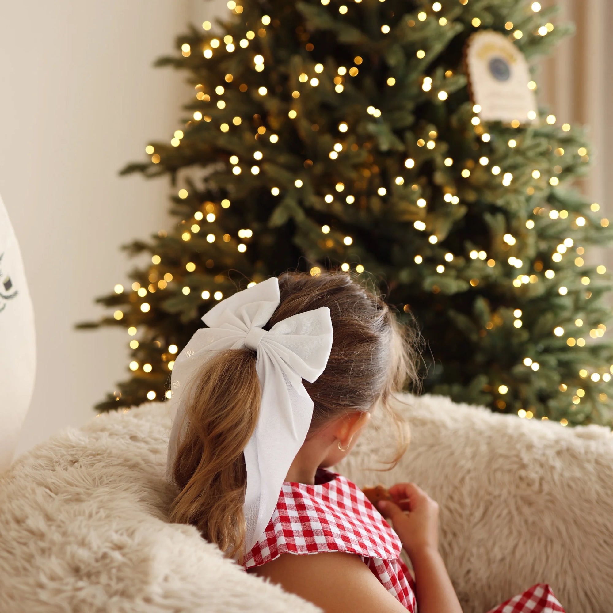 Girl in a red gingham Christmas dress with a large white bow in her hair, sitting on a sheepskin chair facing a lit Christmas tree