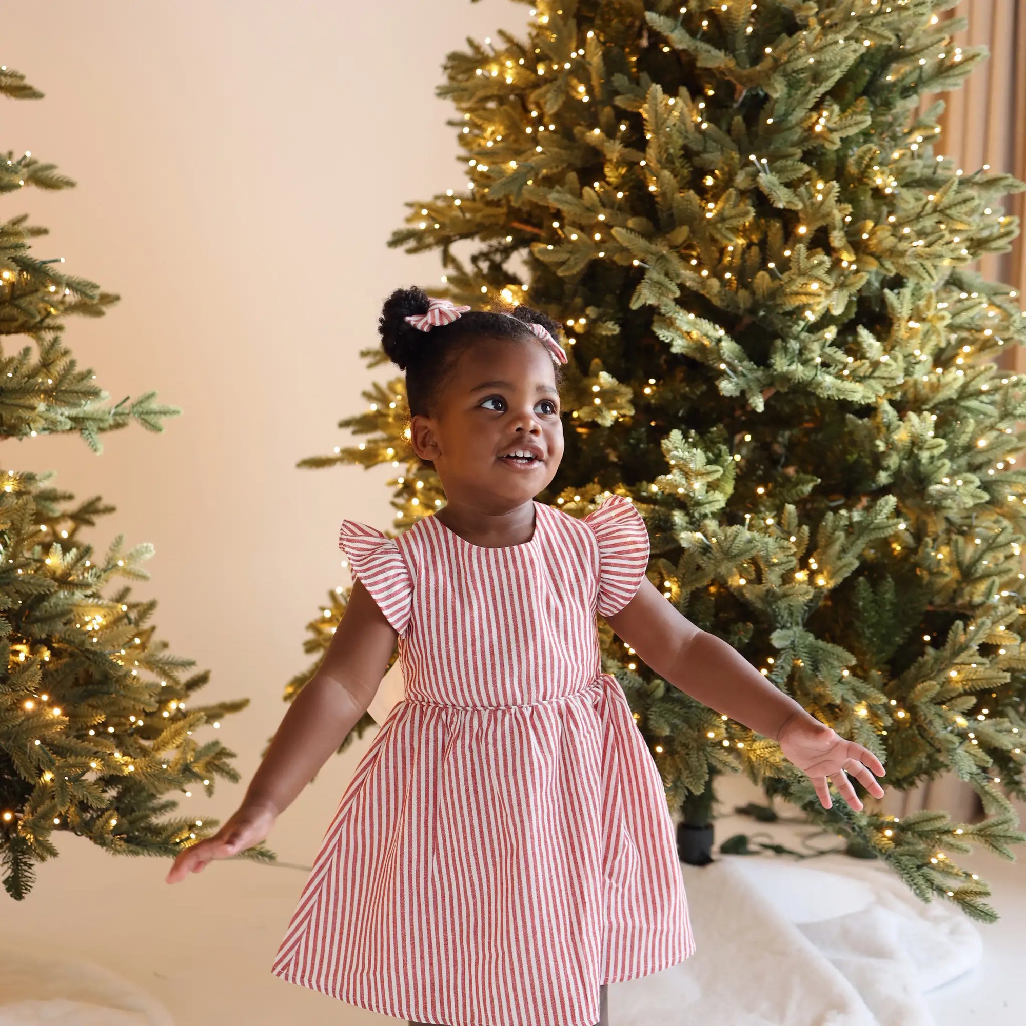Toddler in a red and white stripe flutter sleeve Christmas dress twirling in front of lit Christmas trees