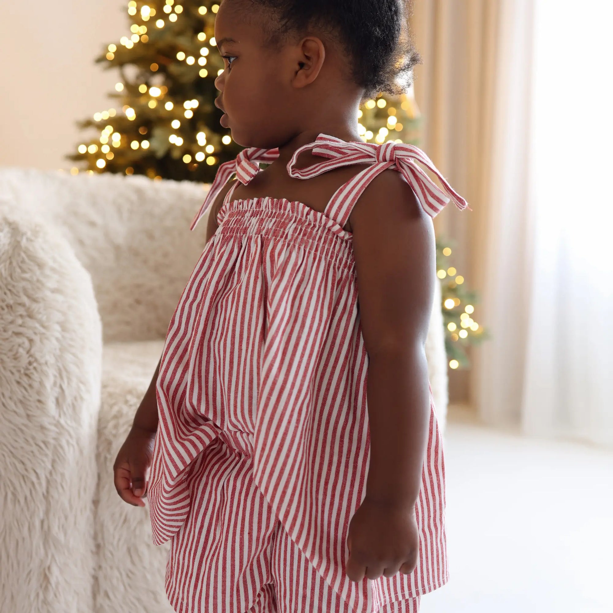 Side profile of a toddler in a red stripe tie shoulder Christmas dress by a lit Christmas tree