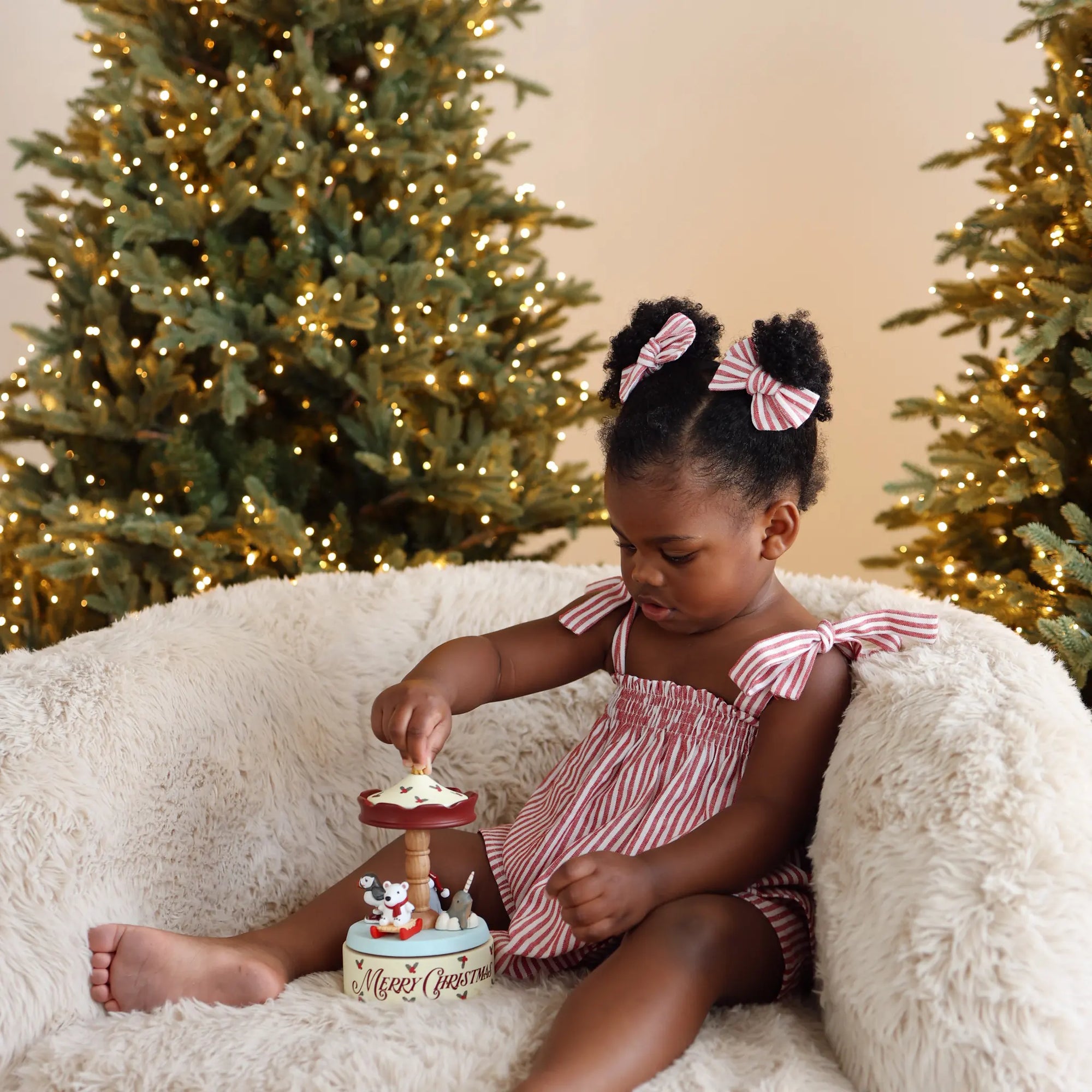 A young girl with her hair in two buns wearing pink and white striped bows sits on a fluffy cream rug, looking down at a small Christmas-themed toy or container. She is wearing a matching pink and white striped romper.