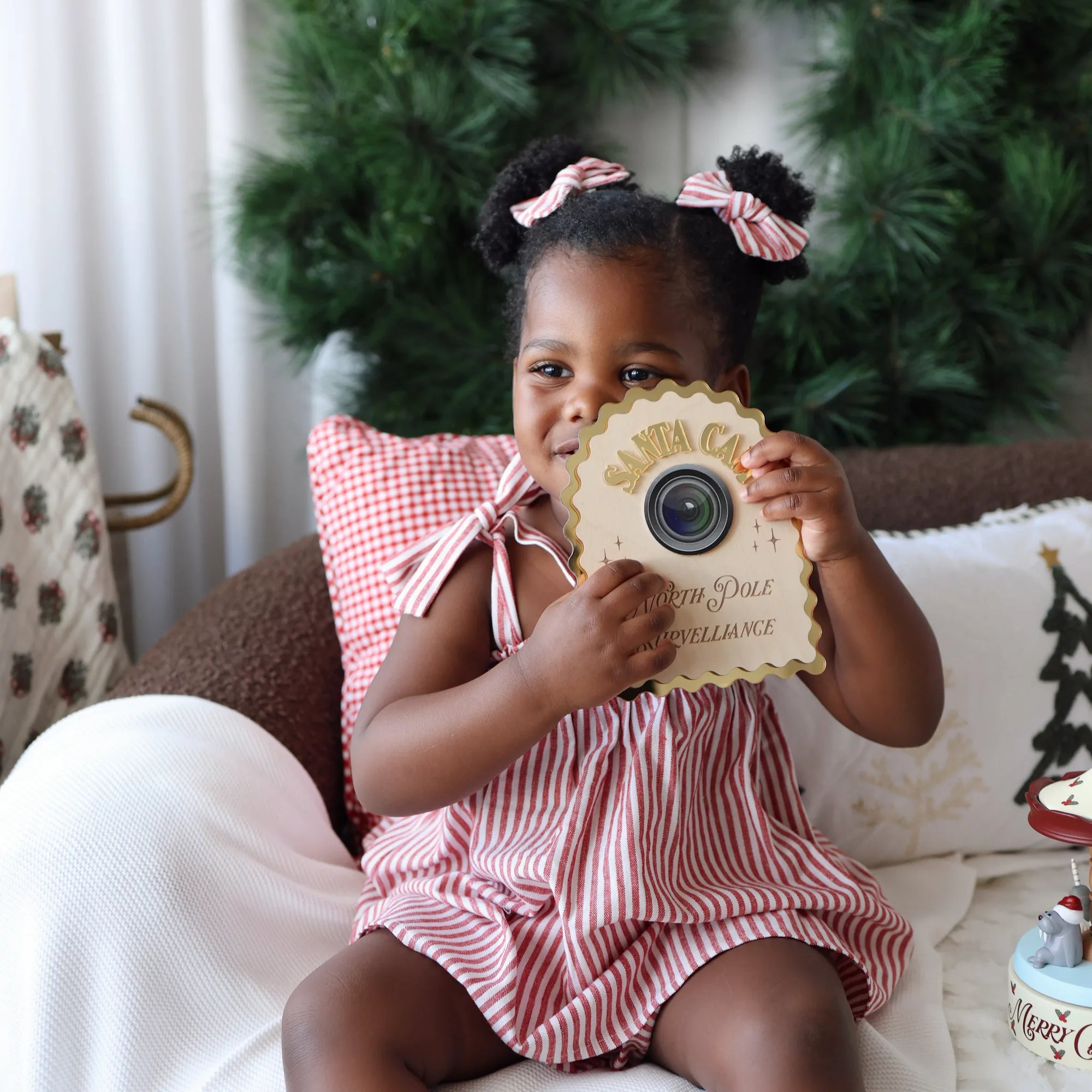 Toddler in a red stripe Christmas dress with matching hair bows holding up a wooden Santa Cam