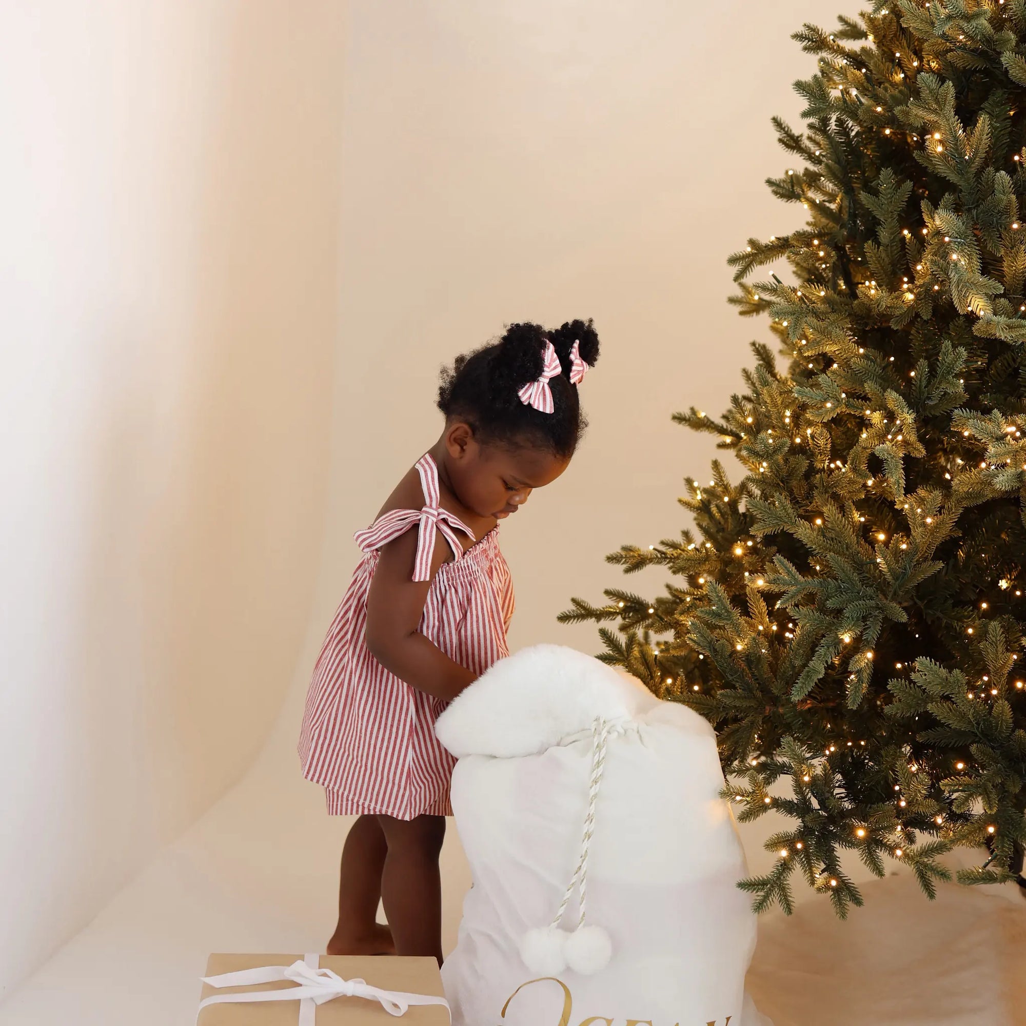 Toddler in a red stripe Christmas dress looking into a personalised white Santa sack by the tree
