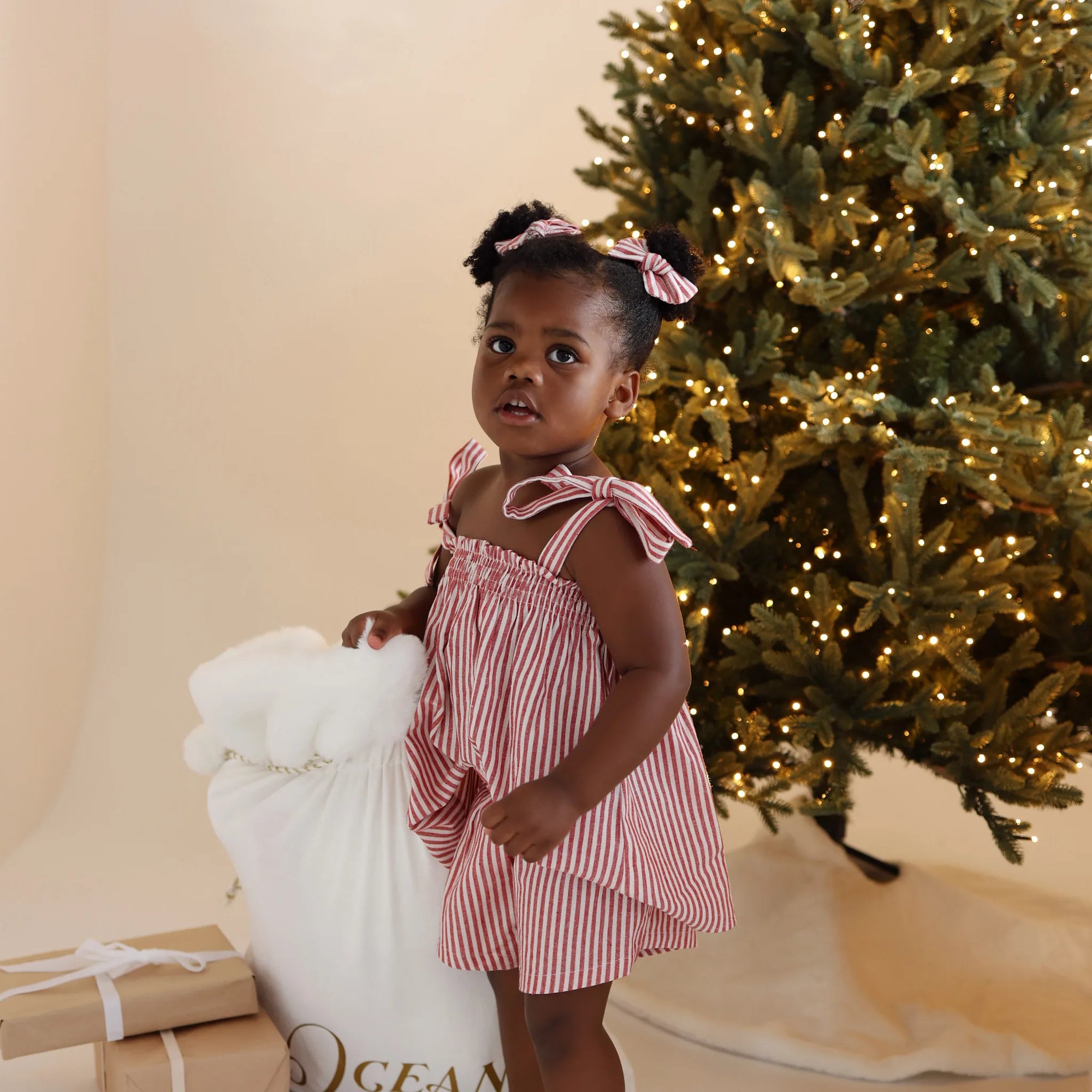 Toddler in a red stripe Christmas dress holding a personalised white Santa sack beside a lit tree