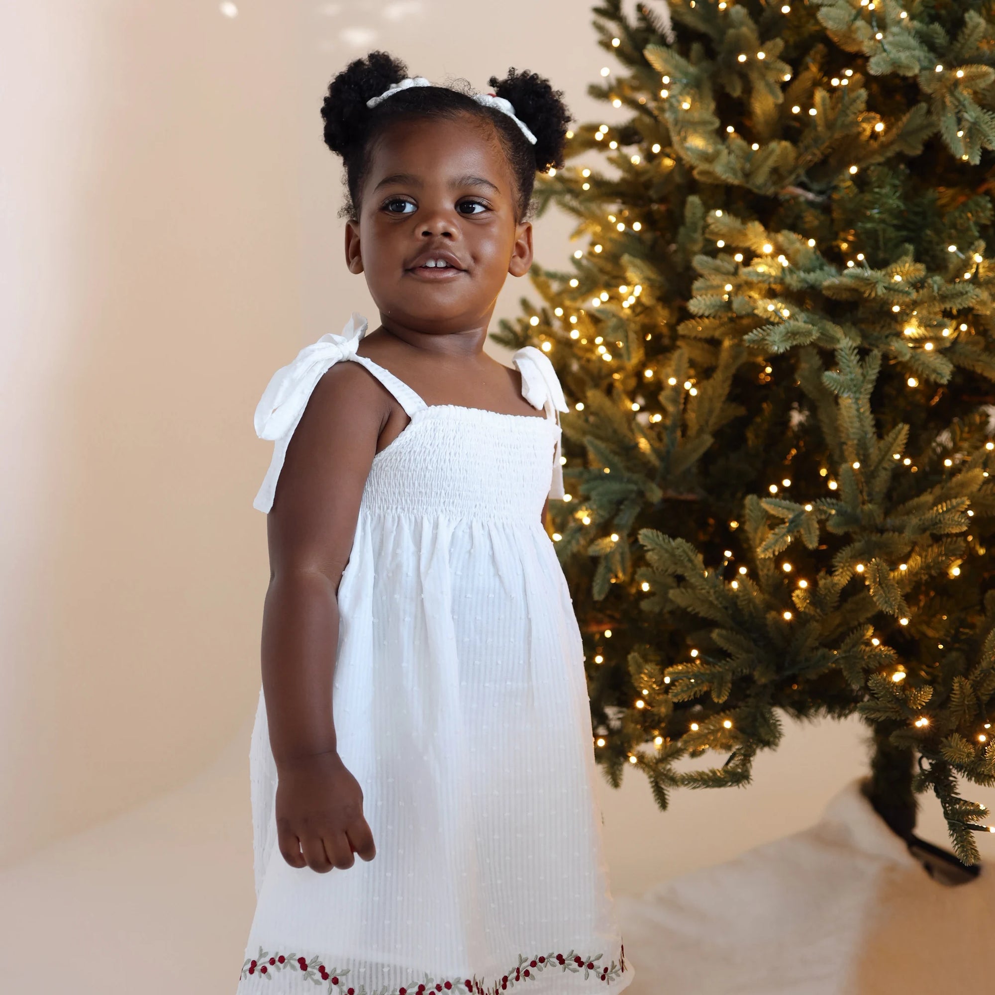 Girl in a white shirred Christmas dress with tie shoulders standing beside a lit Christmas tree