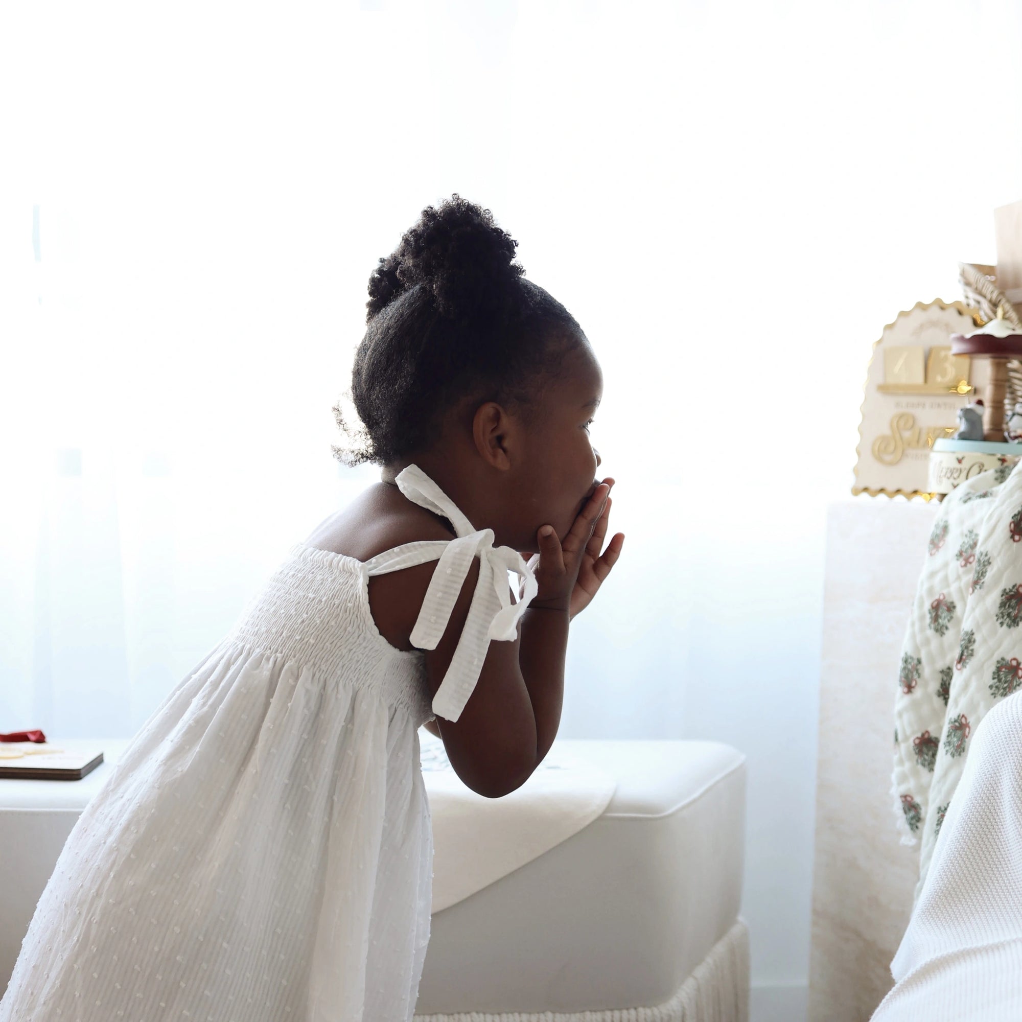 Girl in a white shirred Christmas dress with tie shoulders leaning over a bench by a window