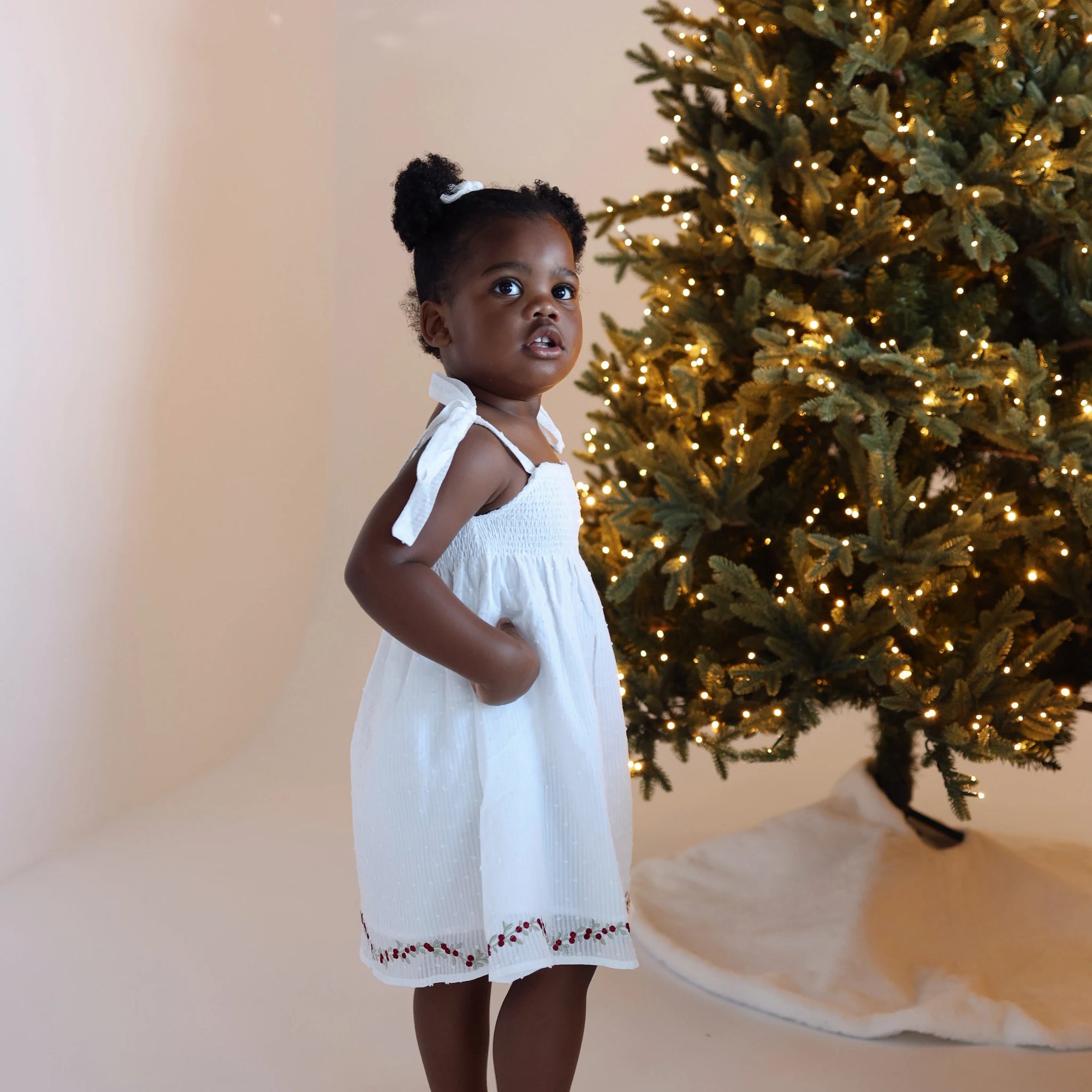 Girl in a white Christmas dress with red embroidered hem looking up beside a lit Christmas tree