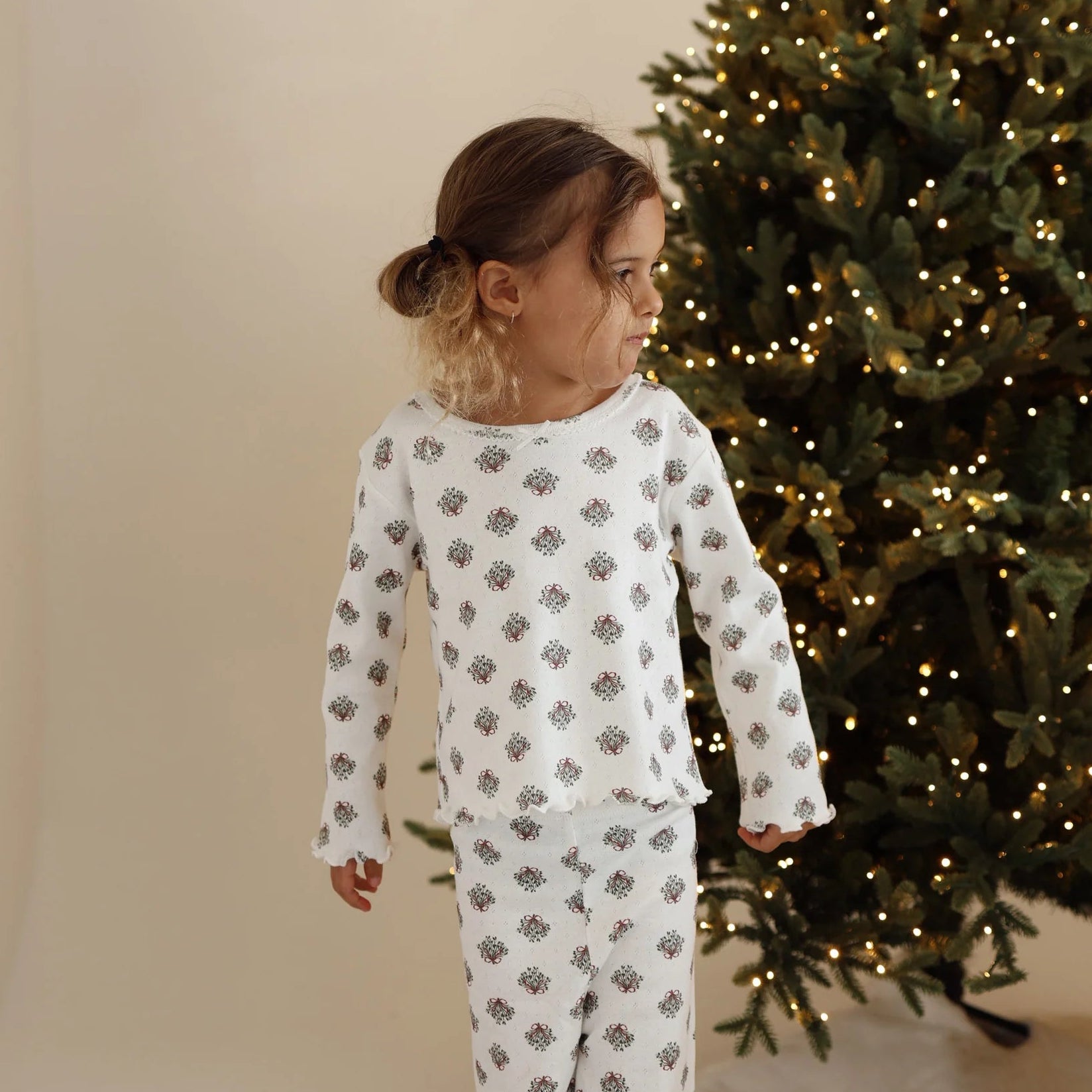 Girl in a wreath print long sleeve pyjama set beside a lit Christmas tree