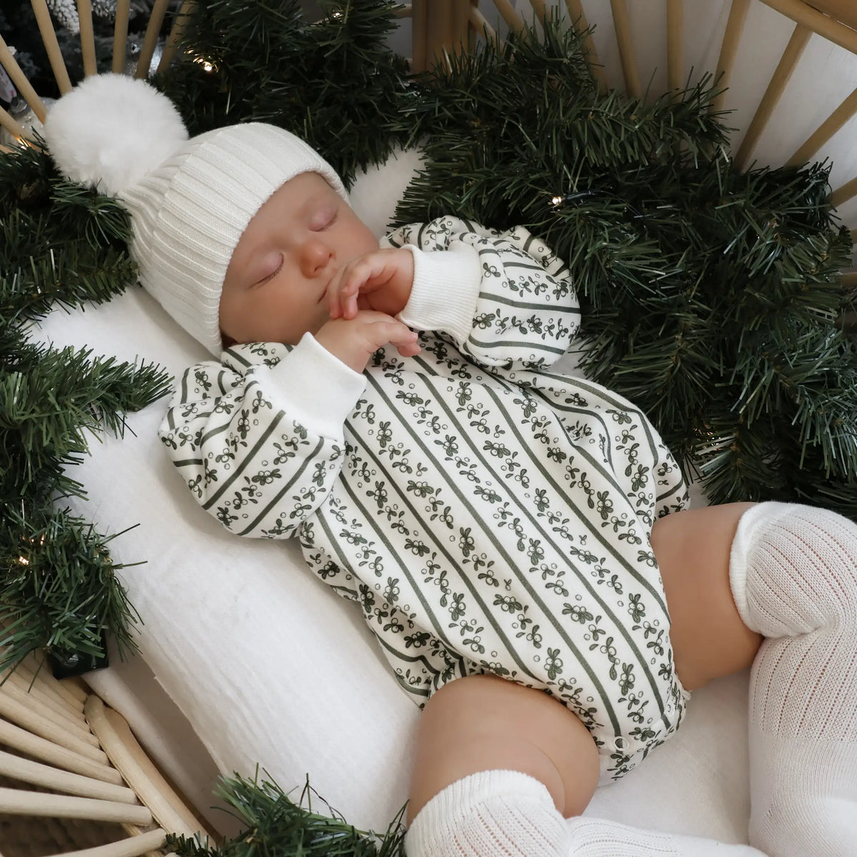 Baby asleep in a holly berry stripe print romper and white pom beanie, nestled in a bassinet surrounded by a pine garland