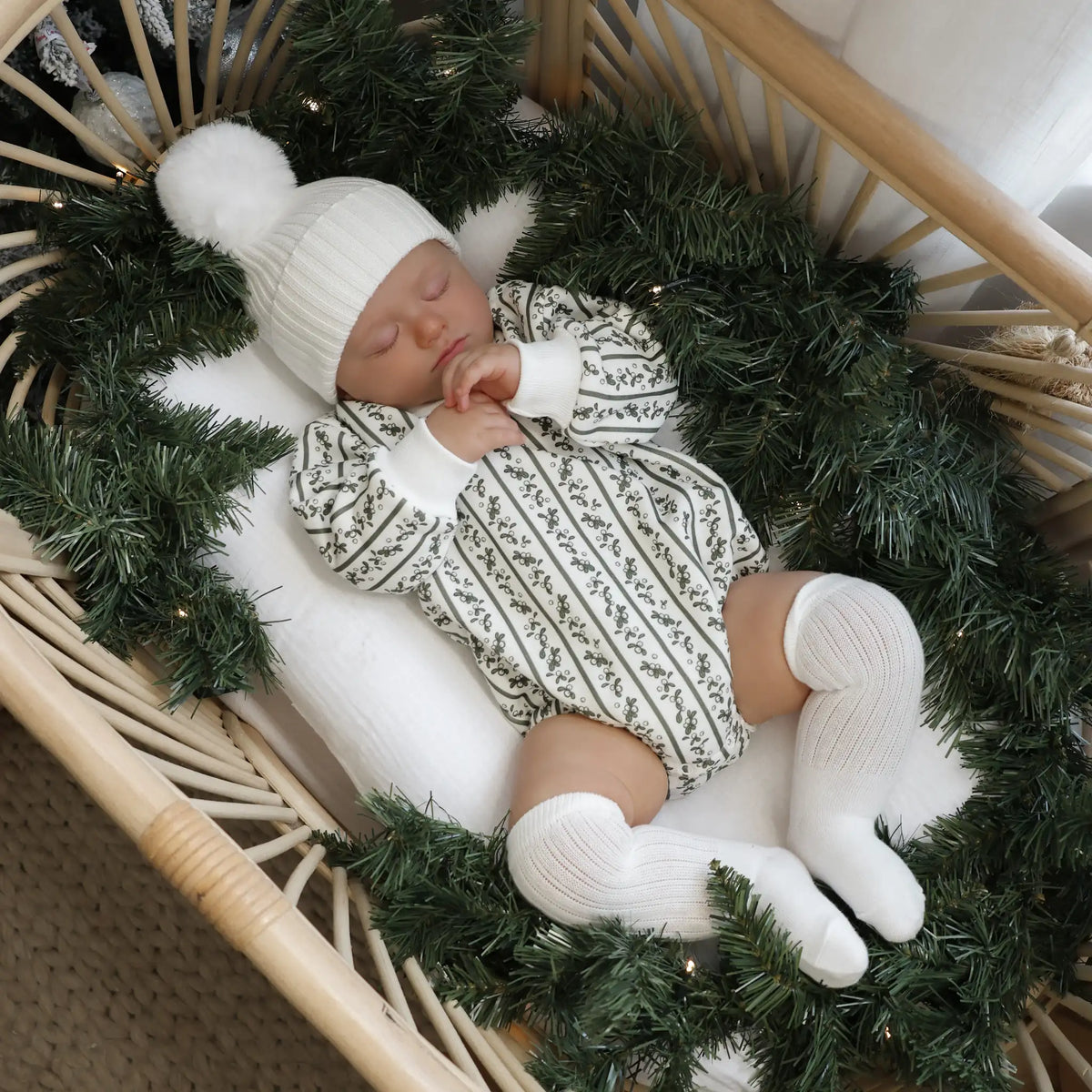 Sleeping baby in a holly berry stripe print romper, white pom beanie and knee high socks, in a rattan bassinet dressed with a pine garland