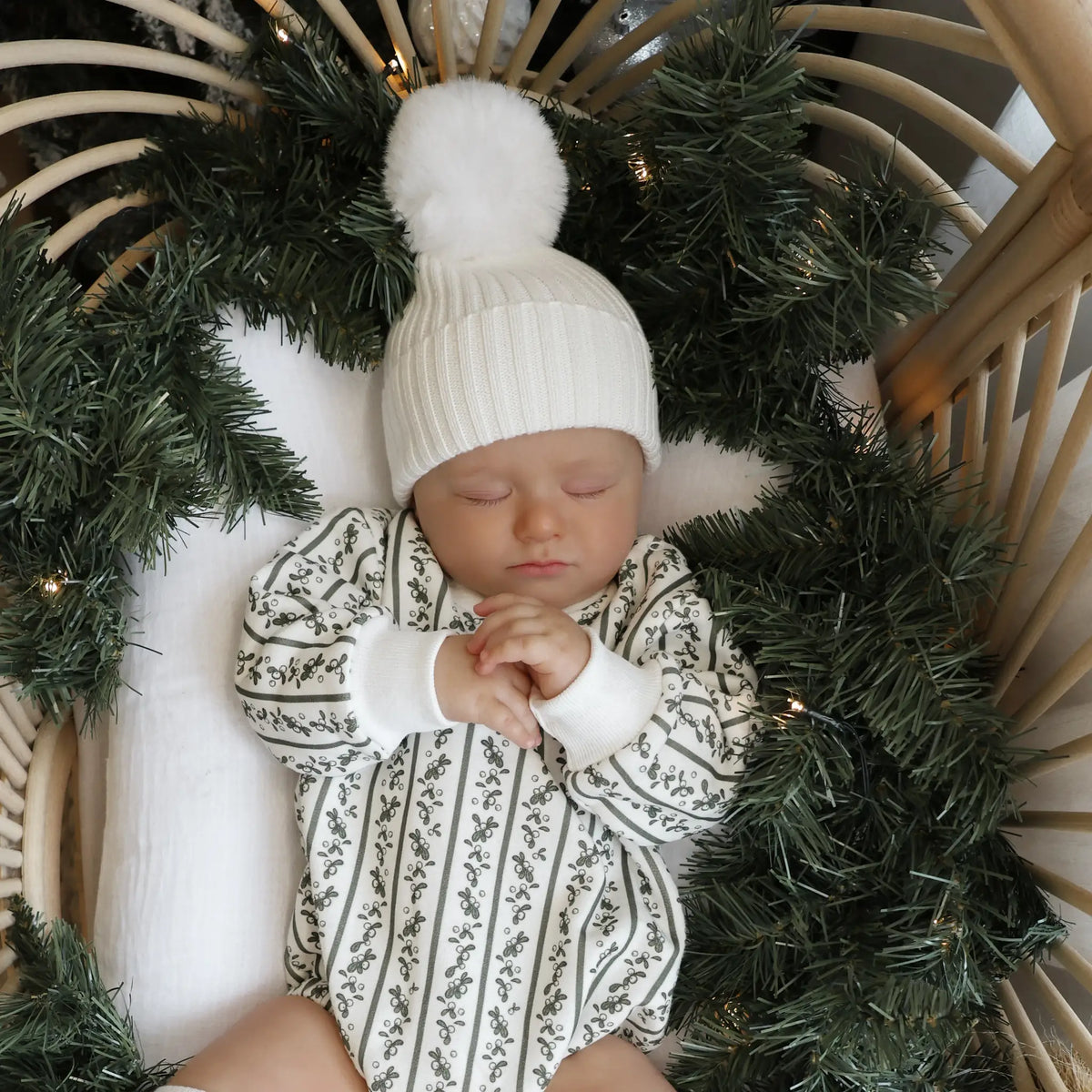 Sleeping baby in a holly berry stripe print romper and white pom beanie, lying in a rattan bassinet framed with a pine garland