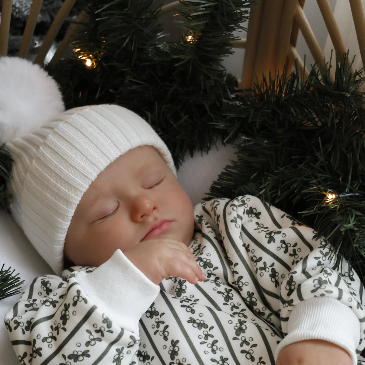 Close-up of a sleeping baby in a holly berry stripe print romper and white pom beanie, surrounded by a lit pine garland