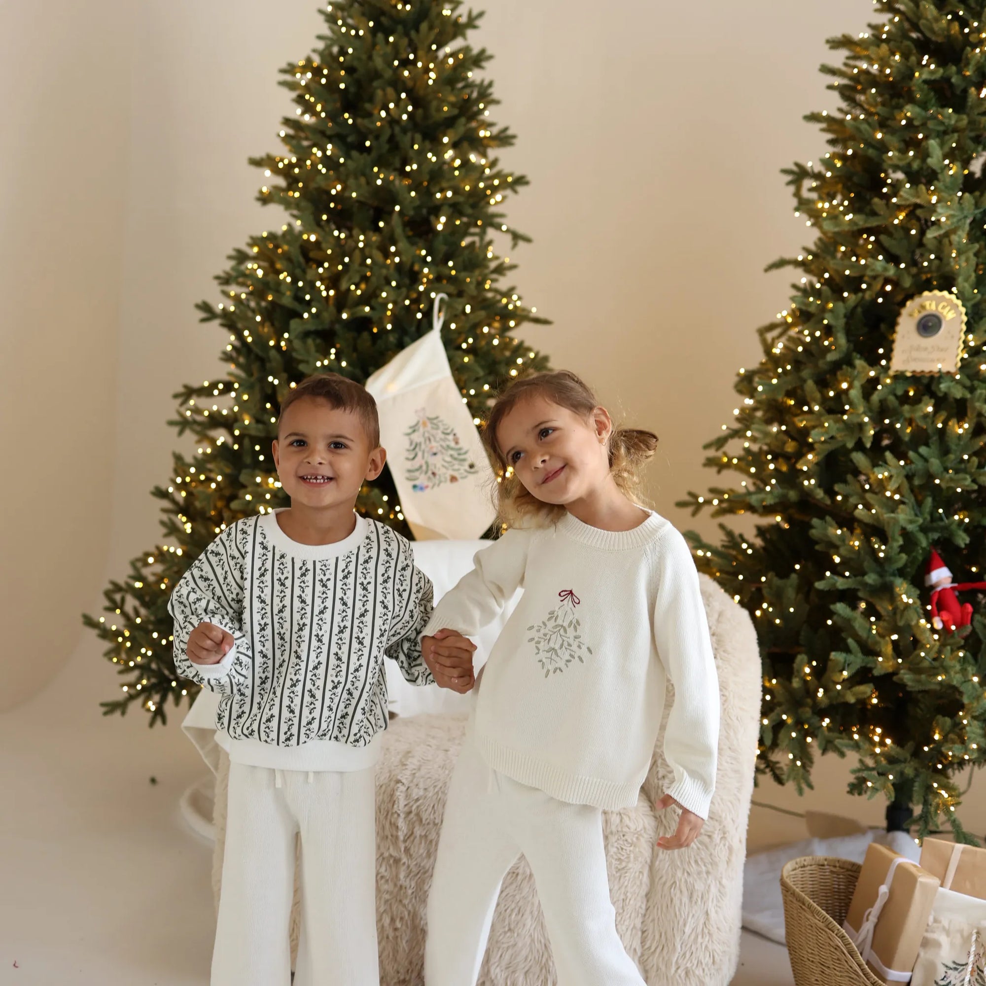 Two children holding hands in cream knit Christmas sets, one in a green botanical print jumper and one with embroidered mistletoe