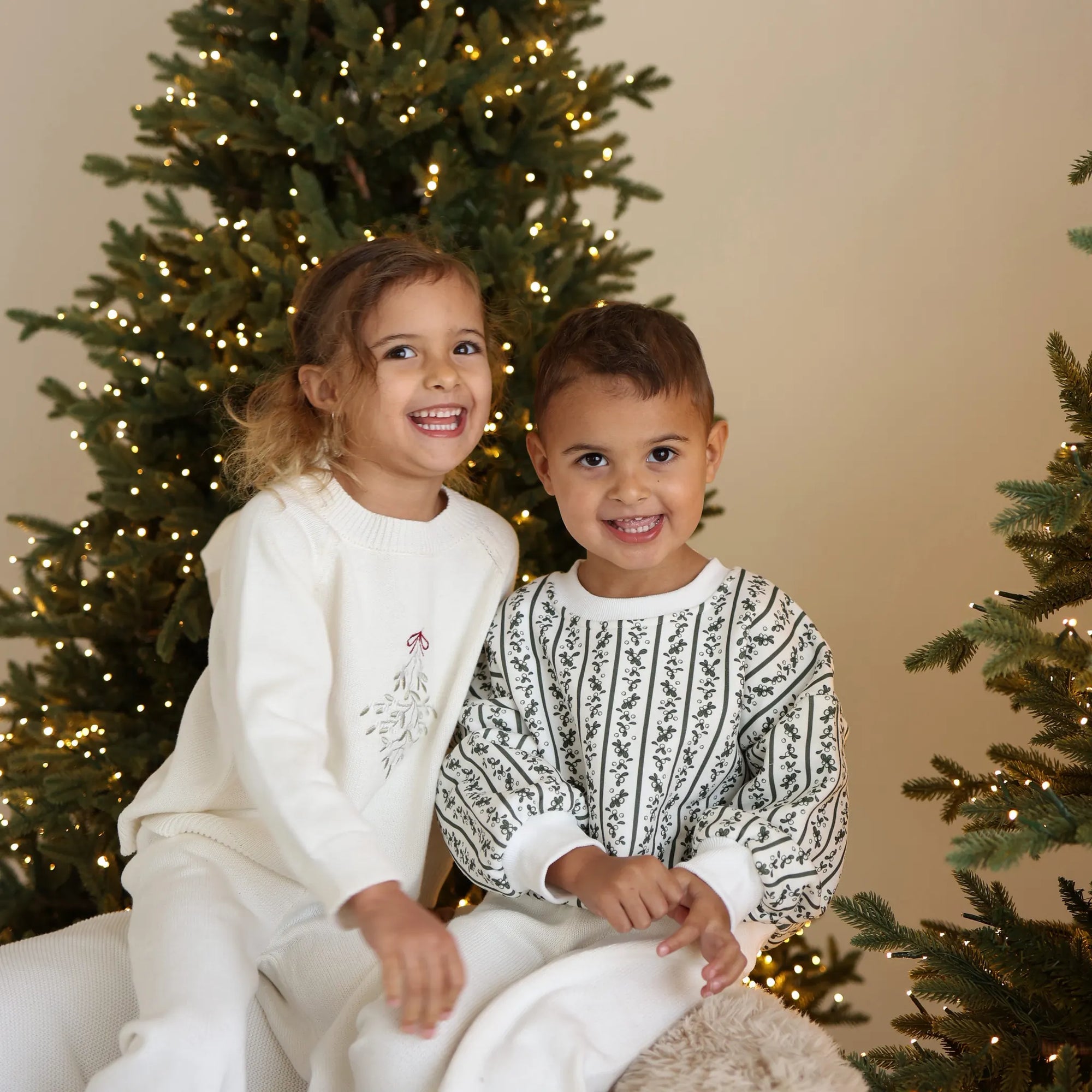 Two children in cream knit Christmas sets sitting together, one with embroidered mistletoe and one in a green botanical print jumper