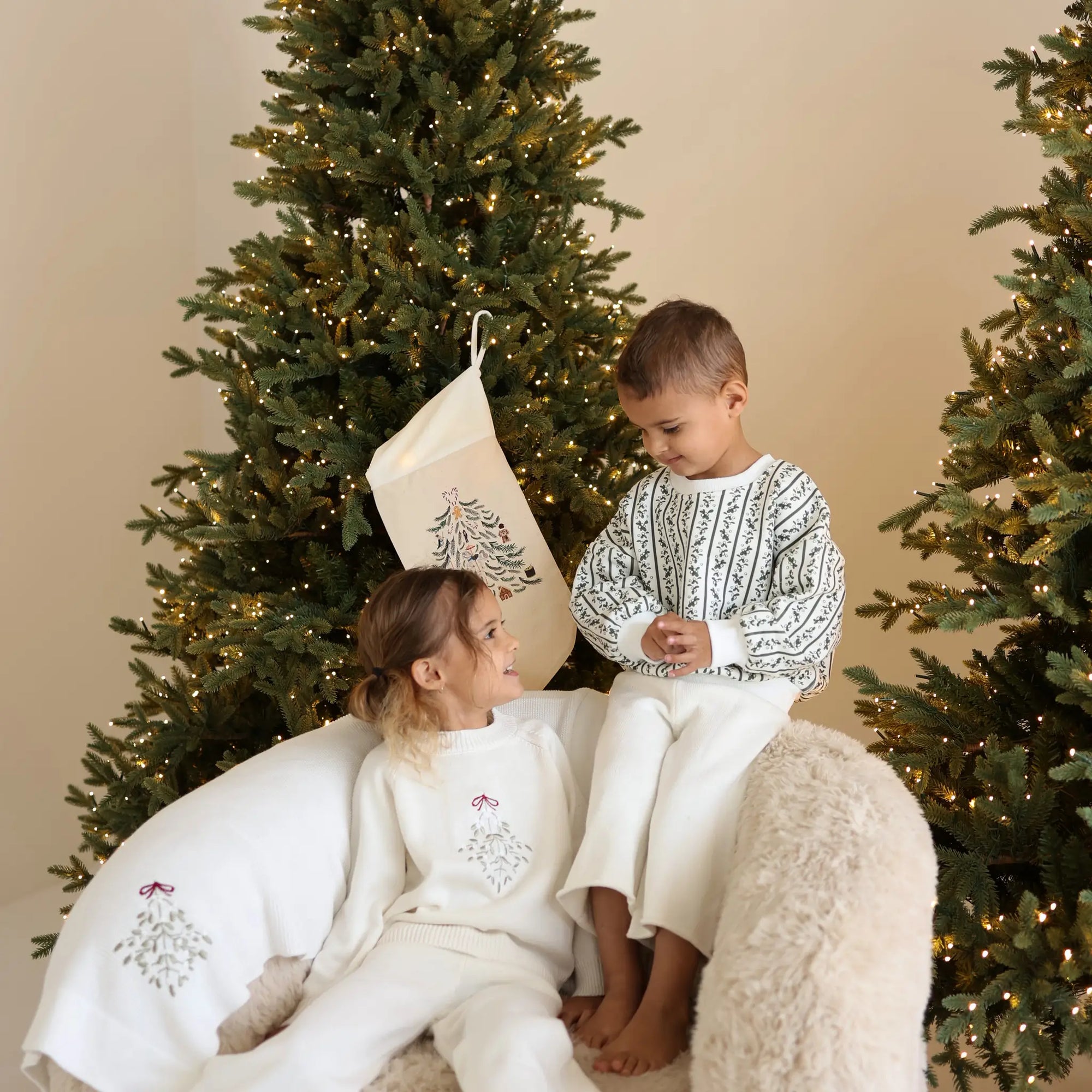 Two children in cream knit Christmas sets on a sheepskin chair beside an embroidered stocking and lit trees