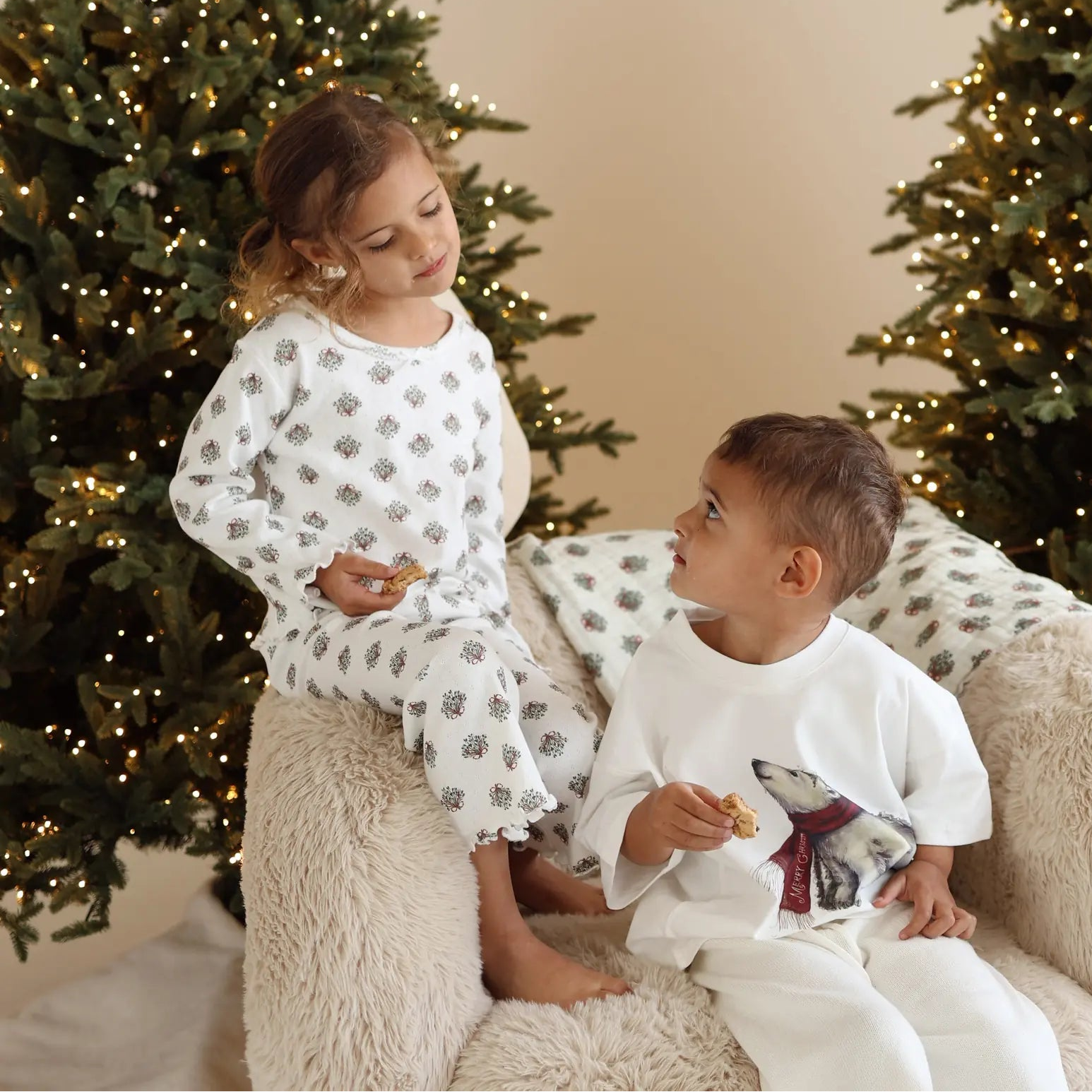 Two children eating cookies on a sheepskin chair, one in wreath print pyjamas and one in a polar bear print tee