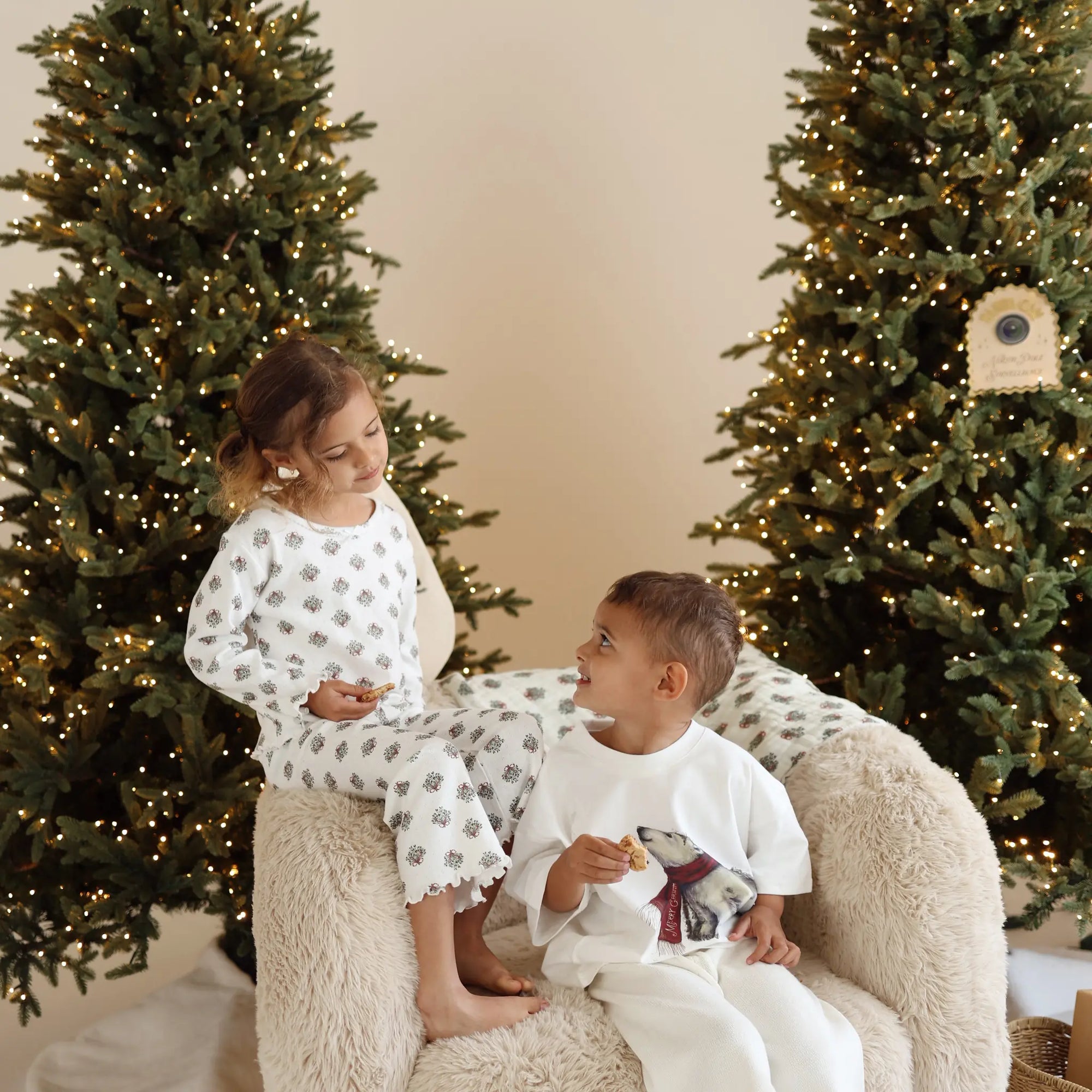 Two children eating cookies on a sheepskin chair, one in wreath print pyjamas and one in a polar bear print tee