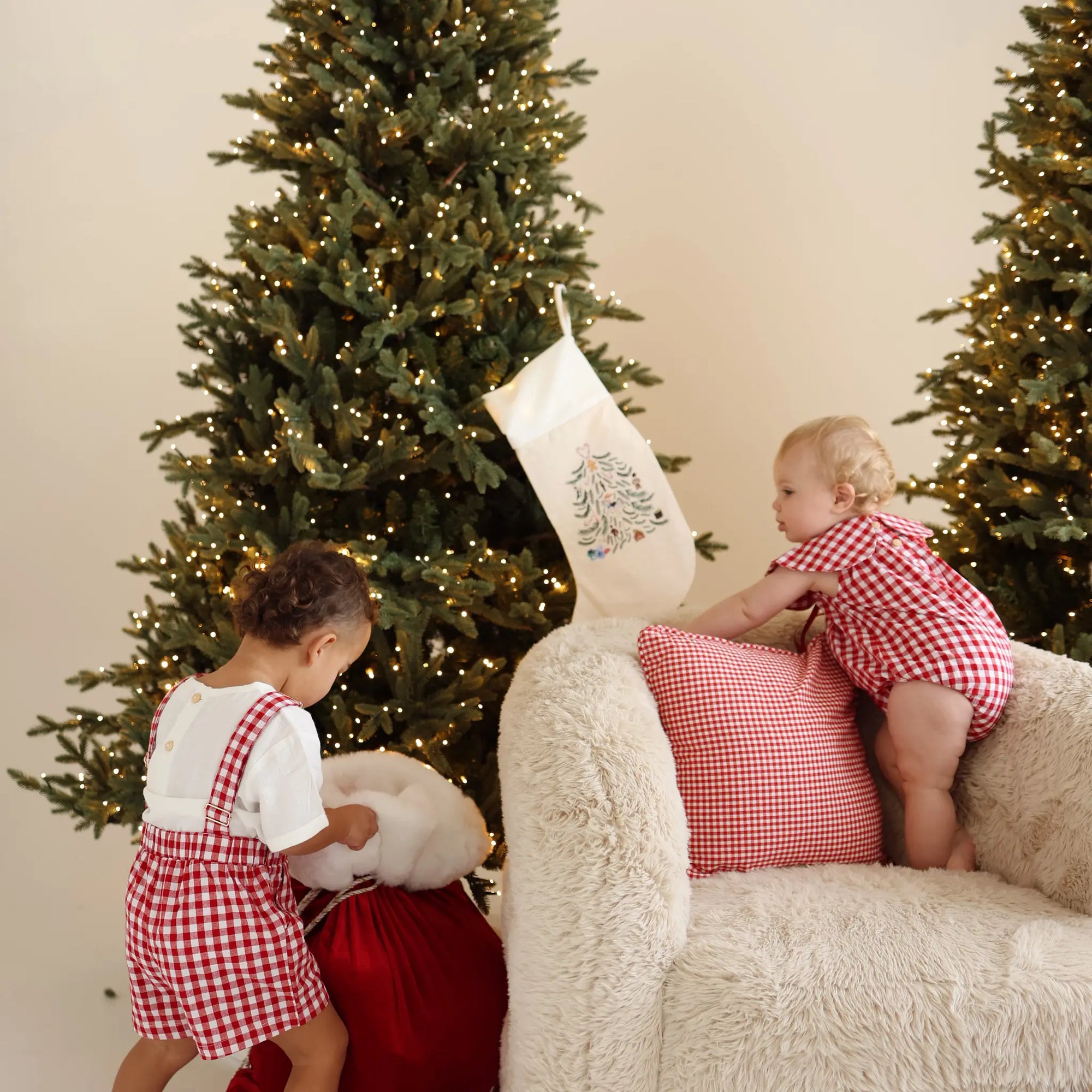 Two children in red gingham Christmas sets by a lit tree, one climbing onto a sheepskin chair
