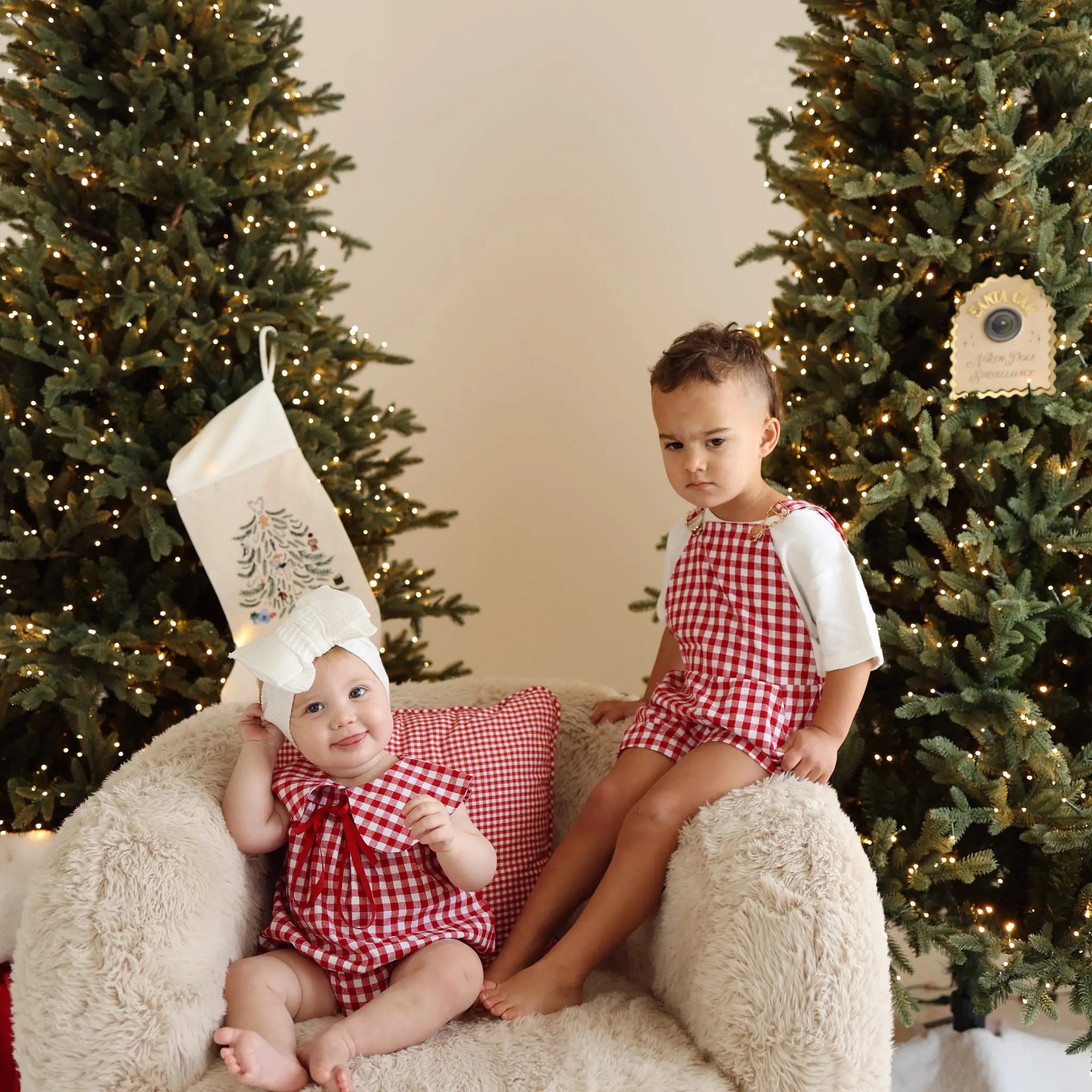 Two children in red gingham Christmas sets on a sheepskin chair, baby holding an embroidered stocking on her head