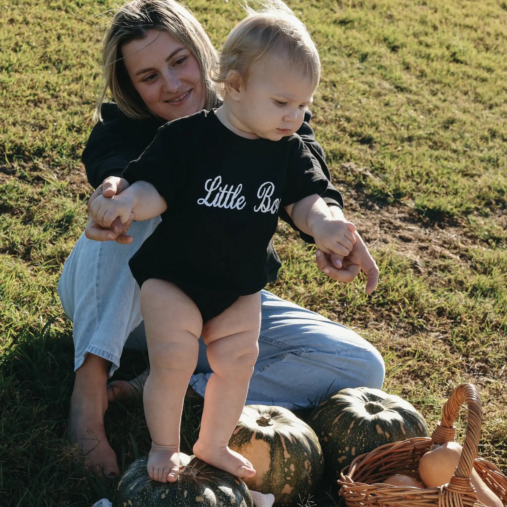 Mum in a Mama Boo sweatshirt steadying a baby in a Little Boo romper standing on pumpkins