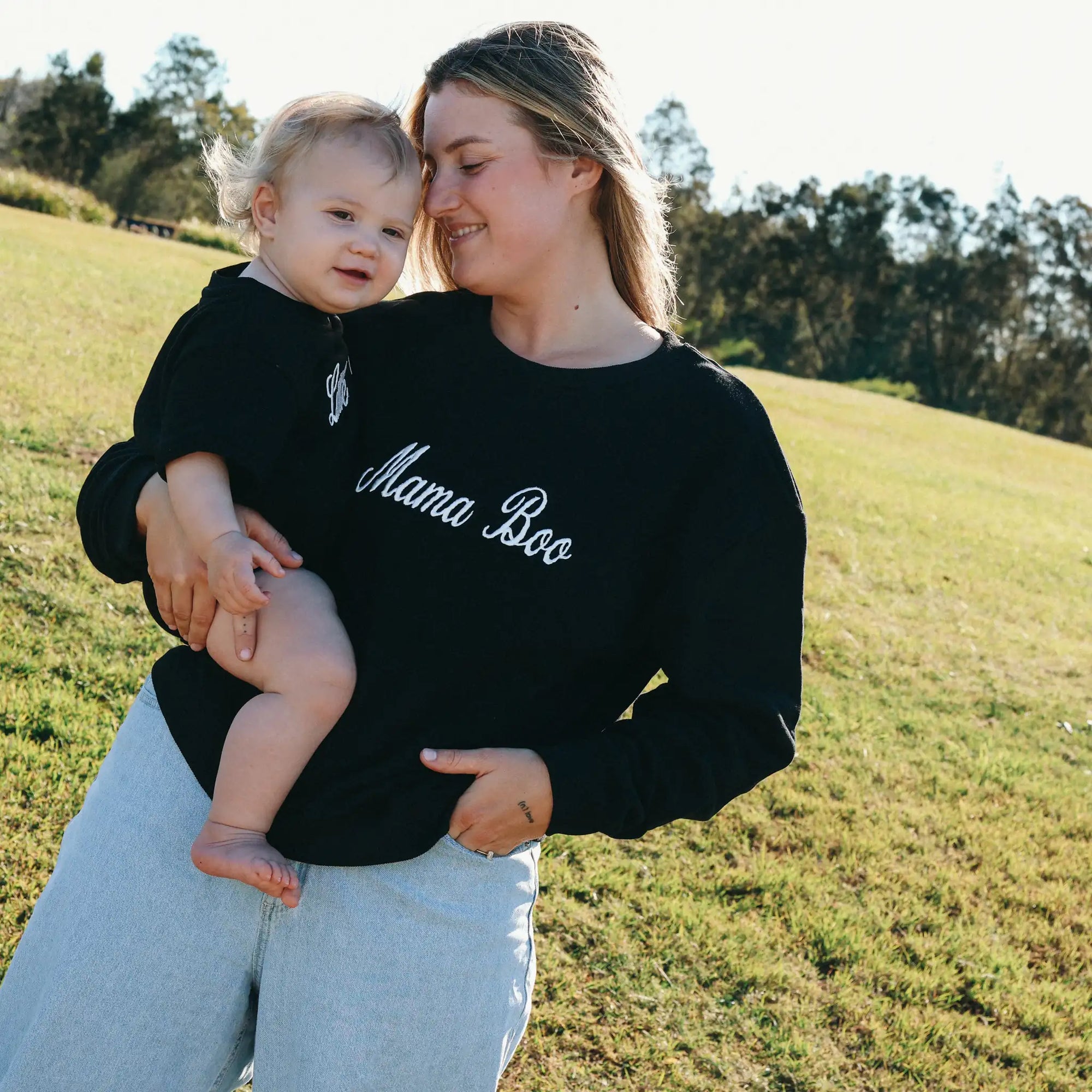 Mum holding her baby in matching black Mama Boo and Little Boo sweatshirts in a sunlit field