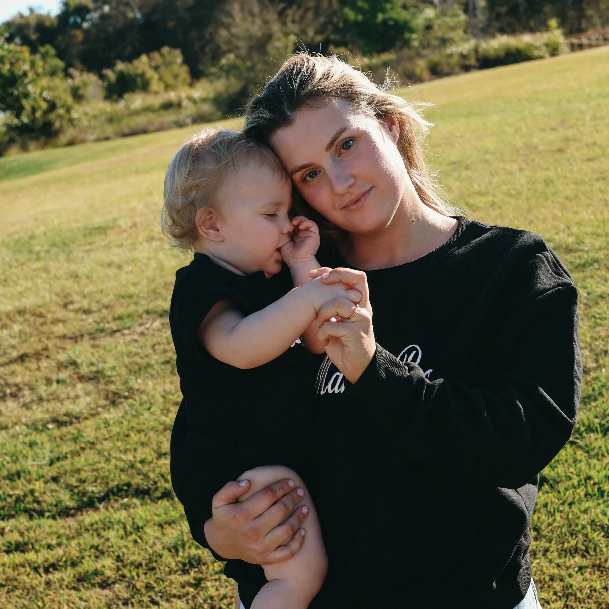 Mum cuddling her baby, both in matching black Mama Boo and Little Boo sweatshirts in a sunlit field