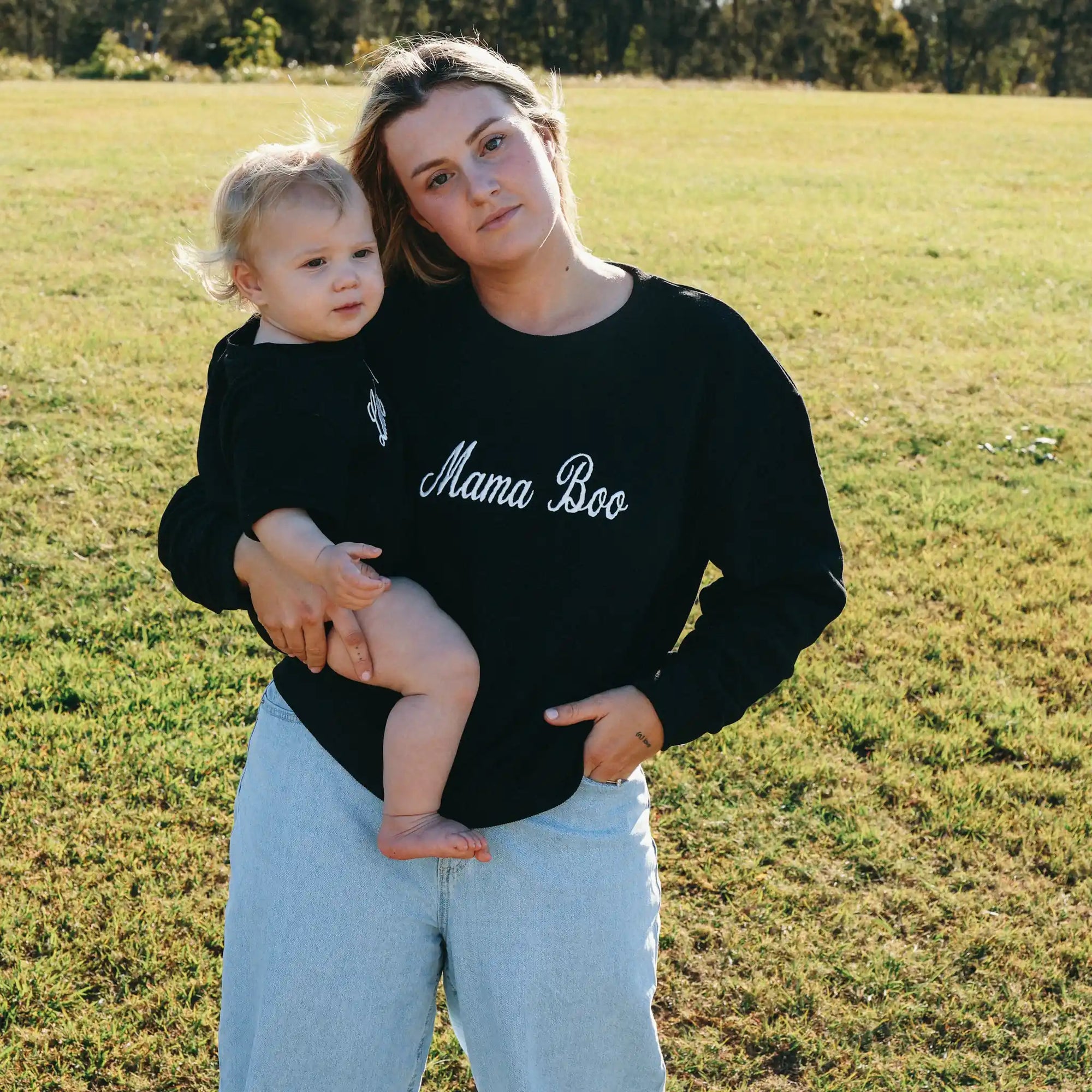 Mum in a Mama Boo embroidered sweatshirt holding a baby in a matching Little Boo sweatshirt in a field