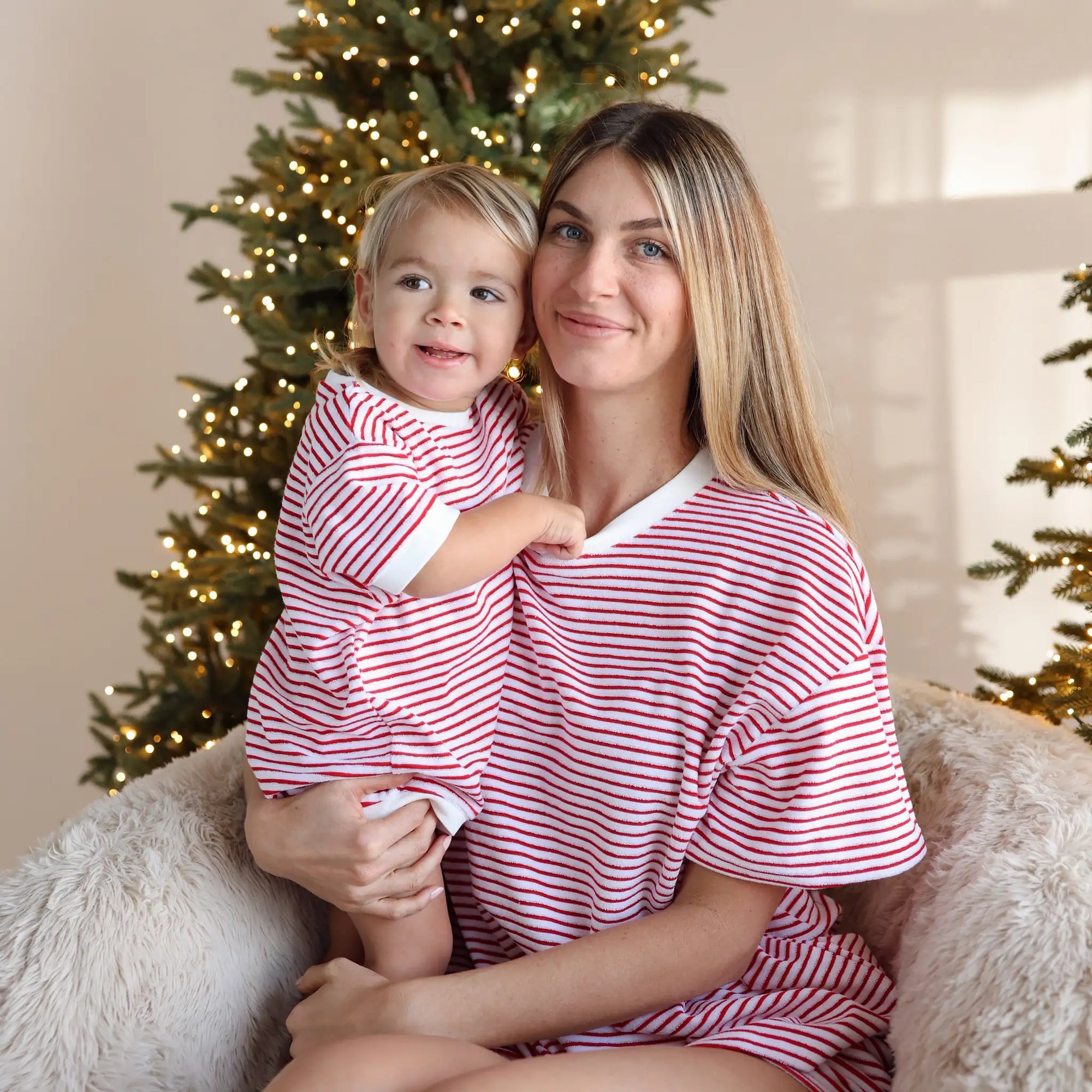 Mum and toddler in matching red and white stripe Christmas pyjamas cuddling in front of a lit Christmas tree
