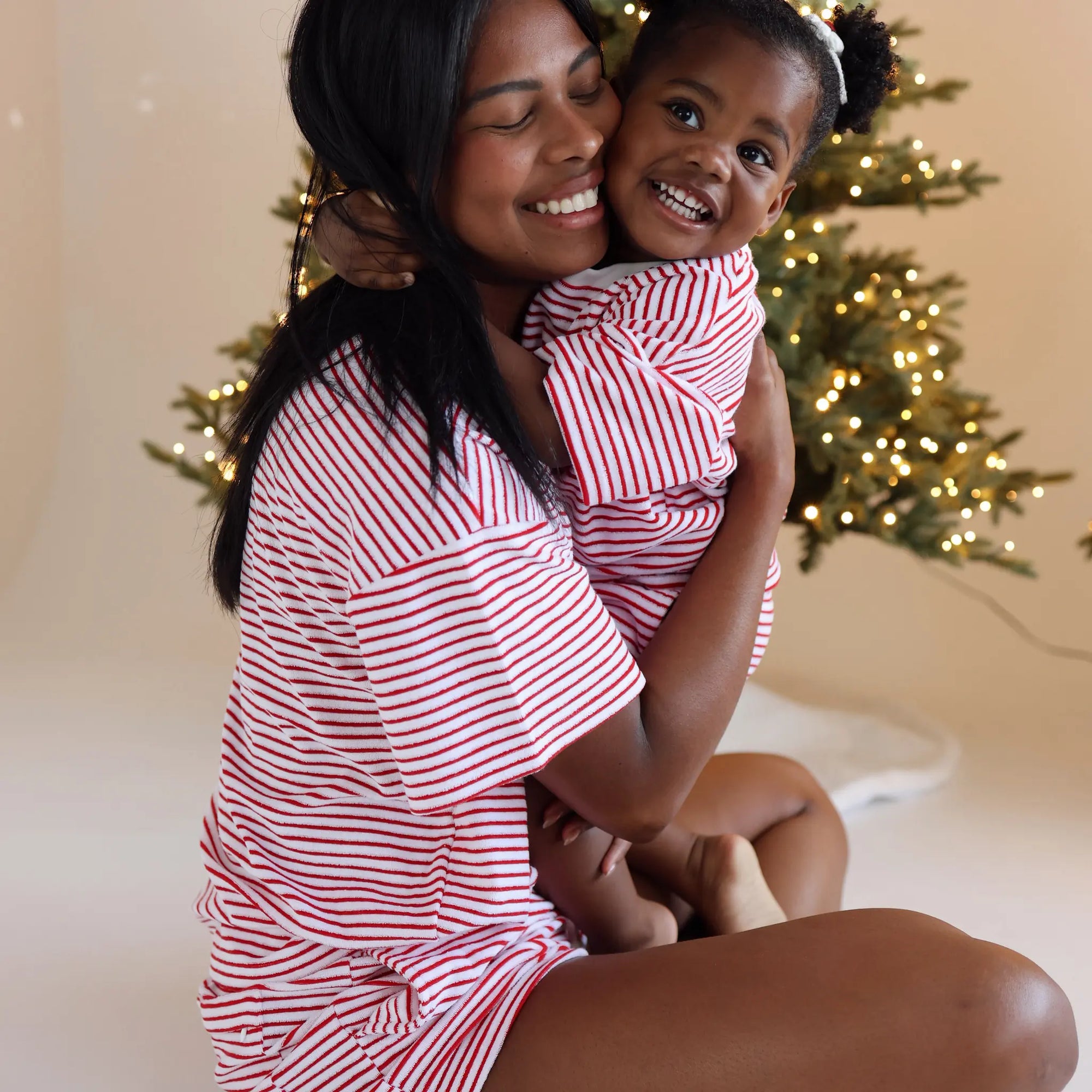 Mum and daughter hugging in matching red and white stripe Christmas pyjamas beside a lit tree