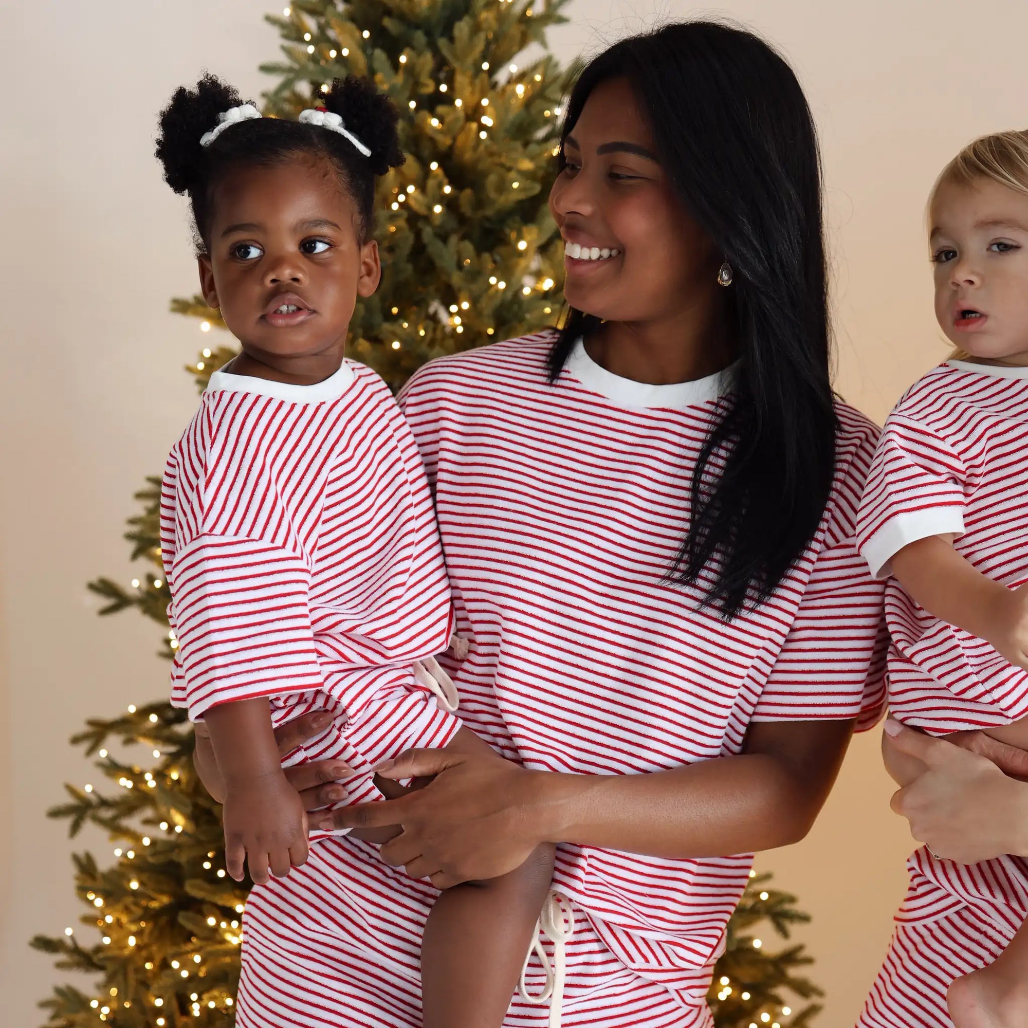 Mum holding two toddlers, all in matching red and white stripe Christmas pyjamas by a lit tree